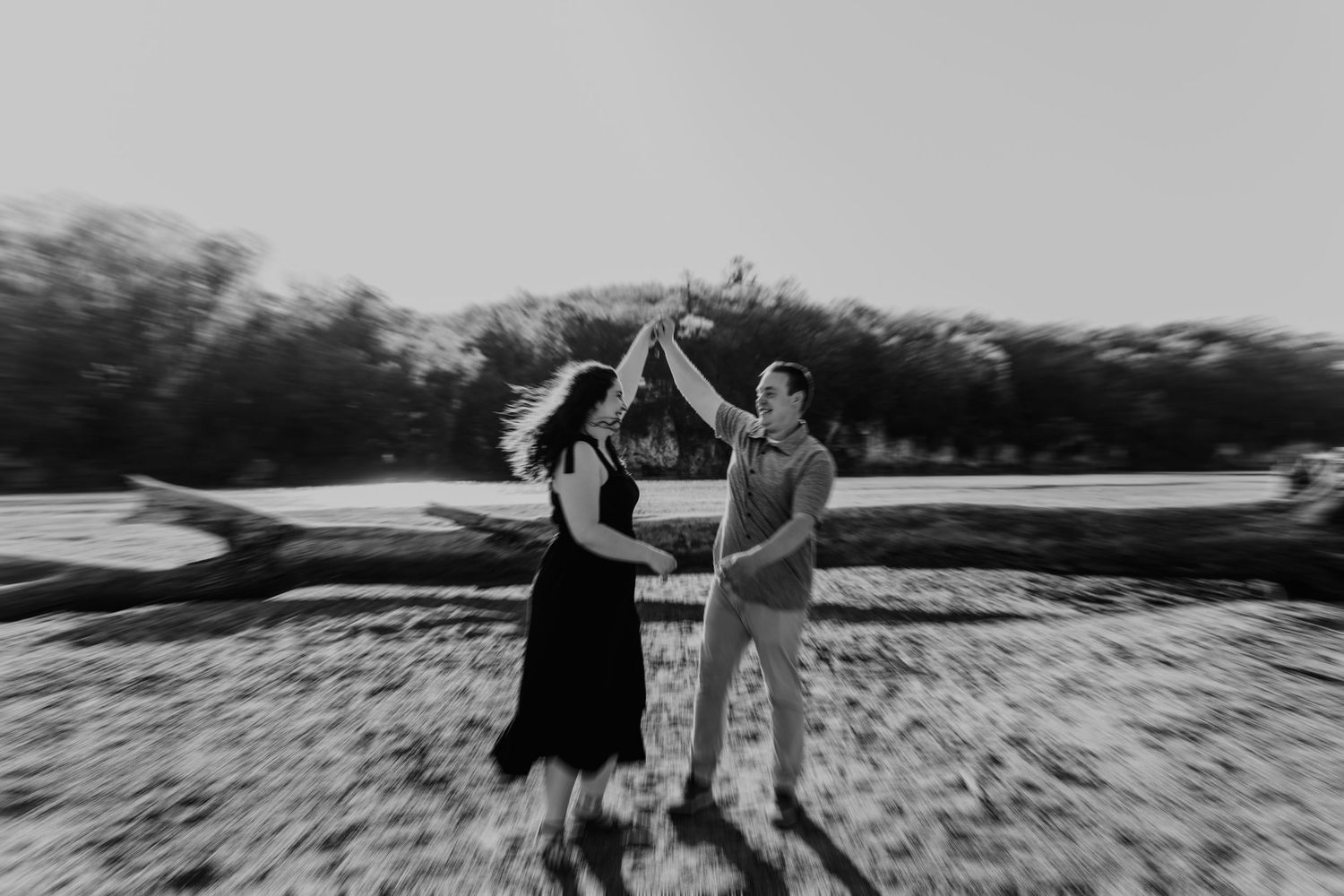 Black and white photo of a couple dancing outdoors, raising hands together joyfully near a fallen log.