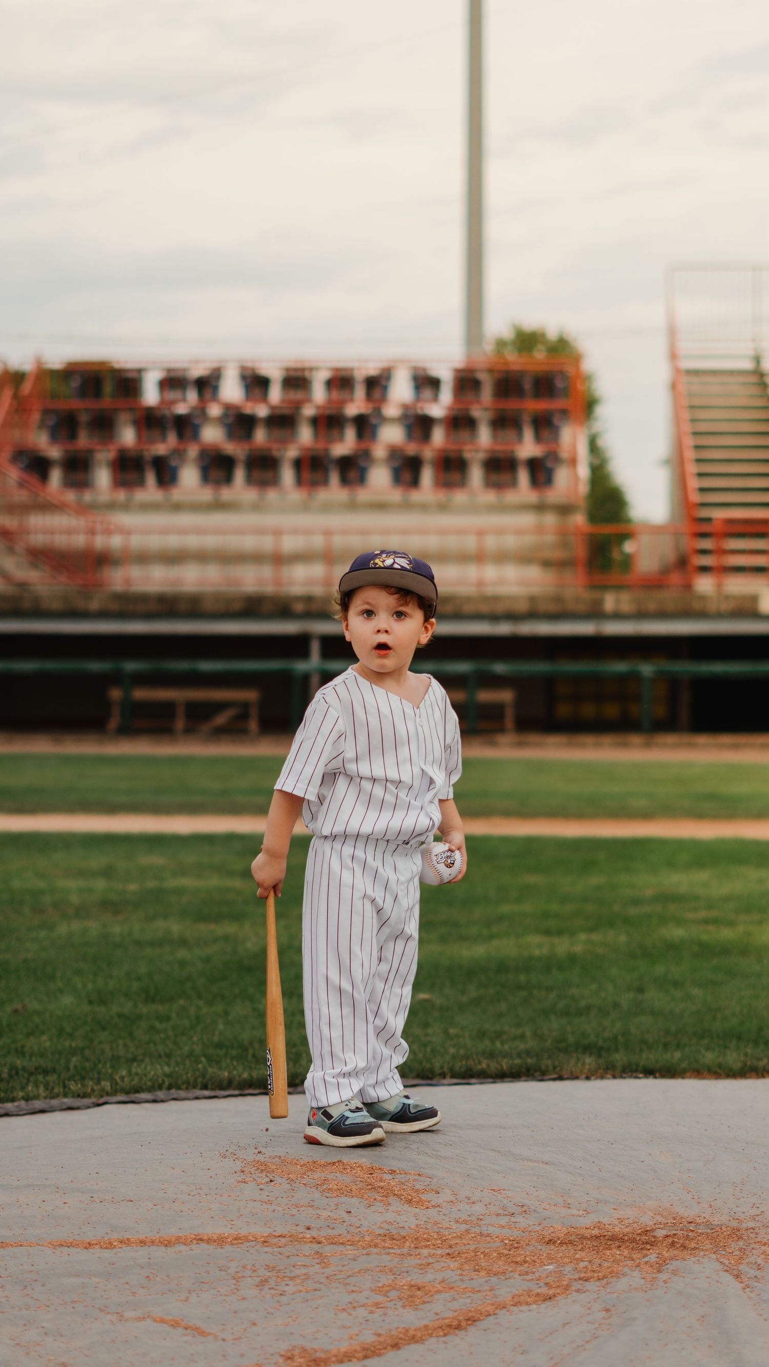 A toddler in a pinstripe baseball uniform holds a bat at a baseball field.