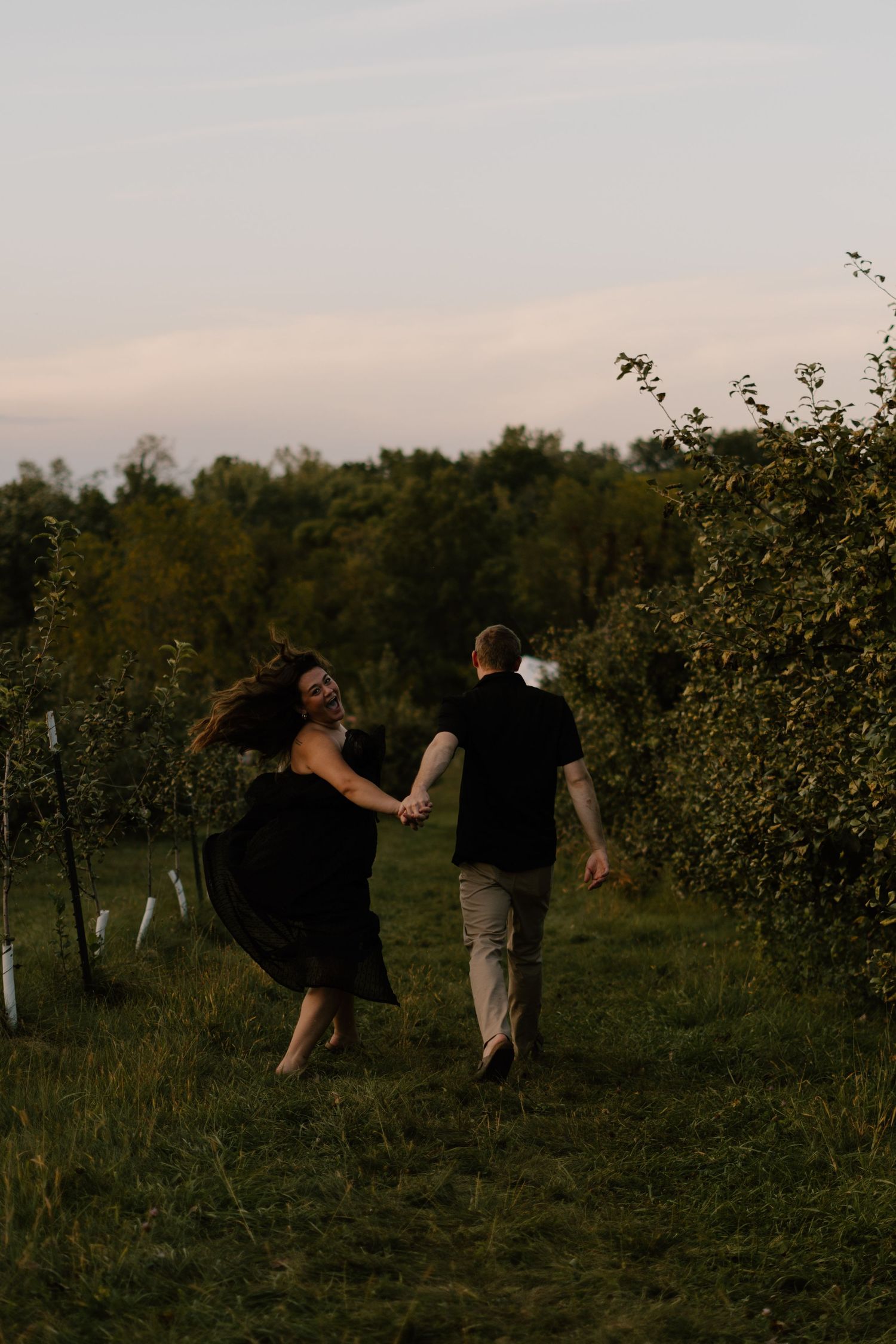 A couple holds hands and runs playfully through a lush green orchard at dusk.