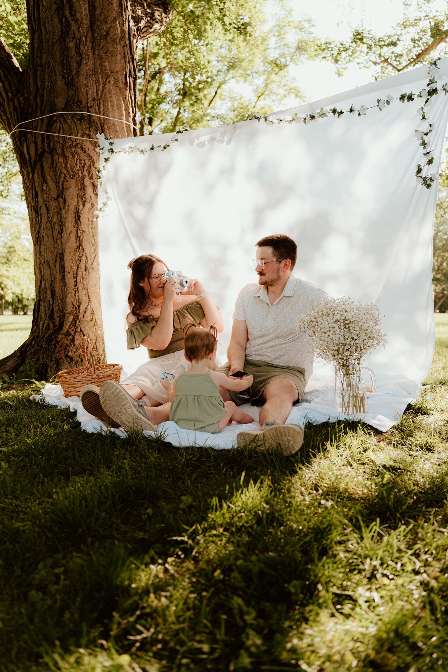 Family enjoying a sunny outdoor picnic on a blanket under a tree with a white backdrop and wildflowers.