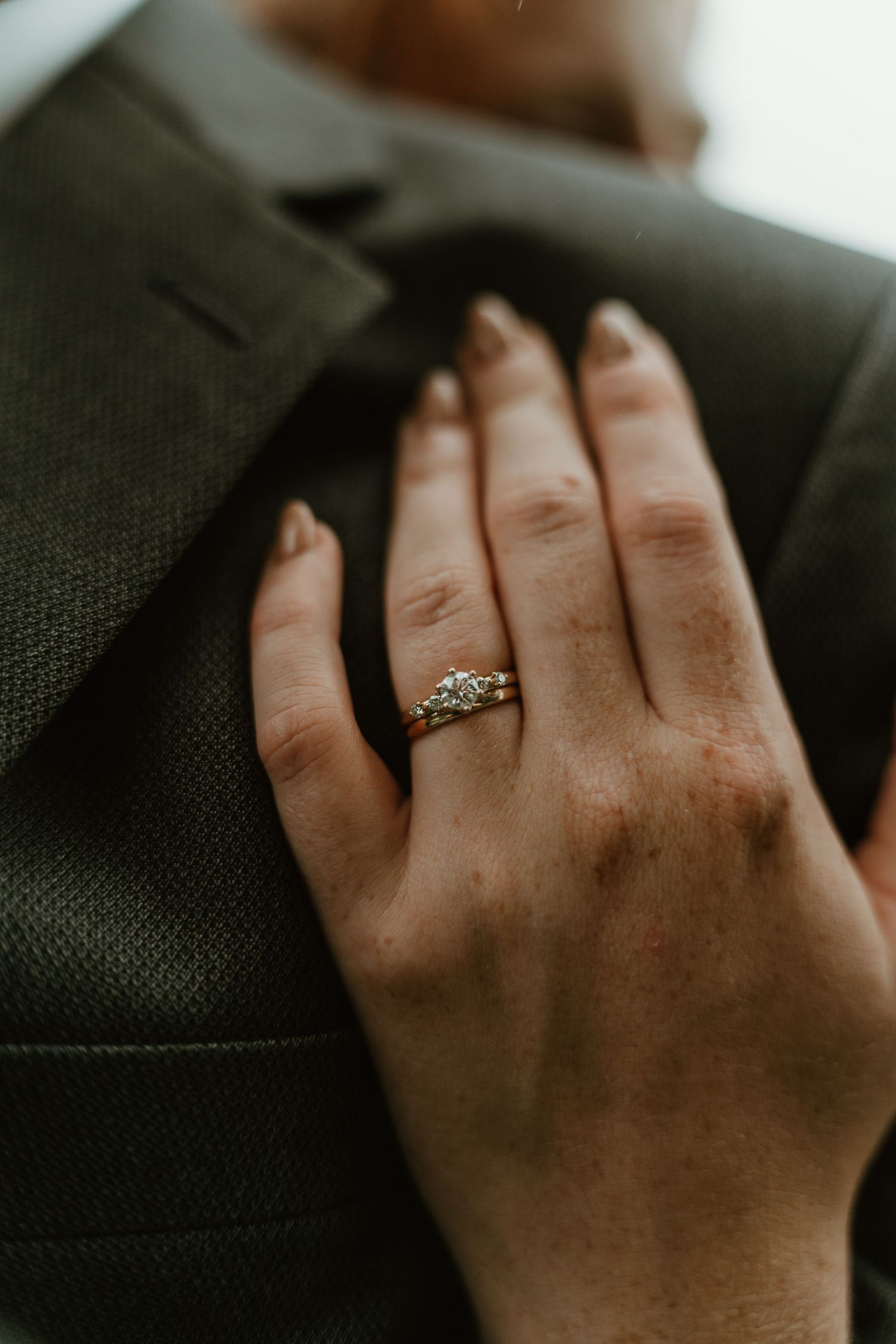 Close-up of a woman's hand displaying a diamond engagement ring and wedding band resting on a dark suit jacket.