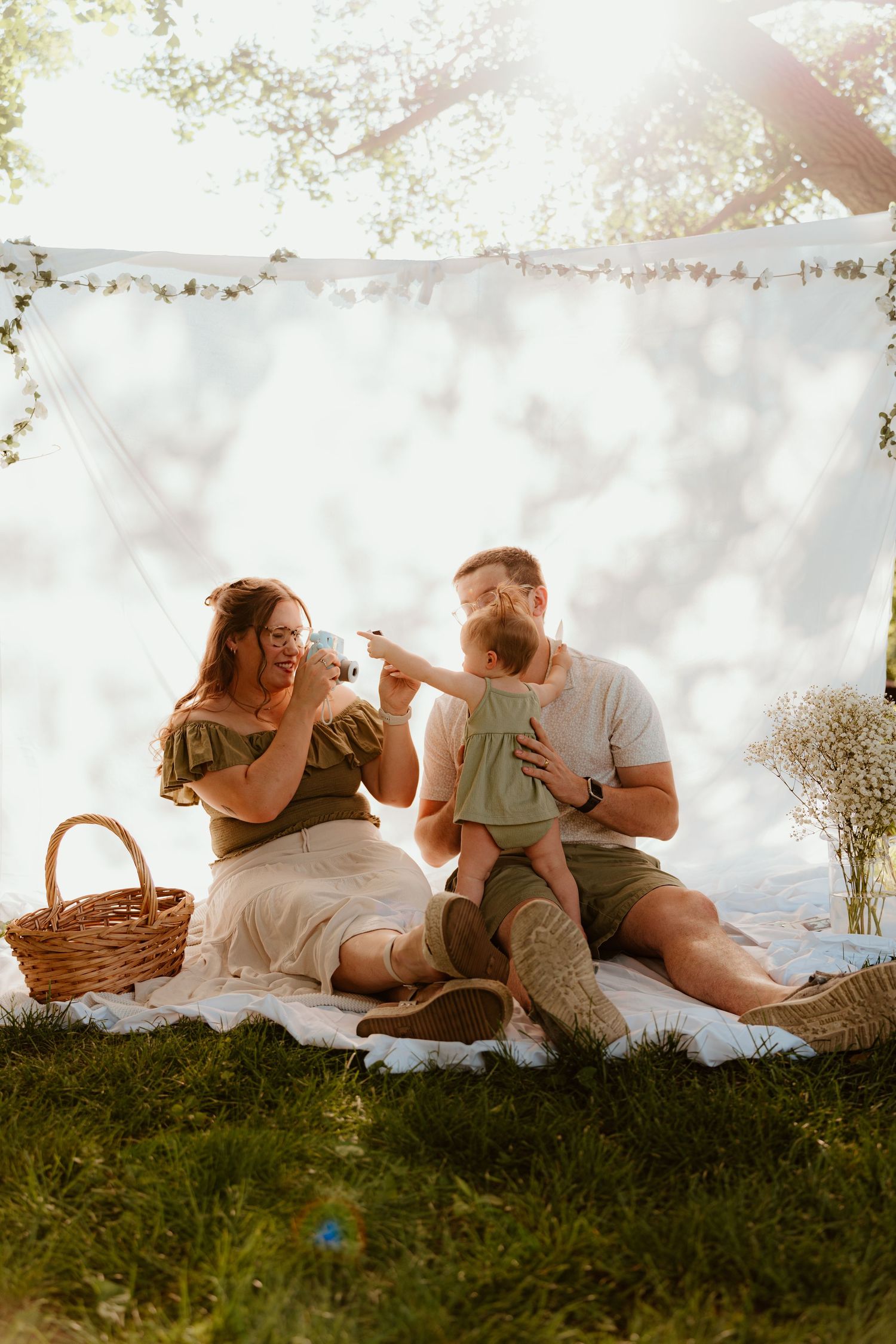 A happy family enjoys a sunny outdoor picnic on a blanket, with parents playing with their baby near a white backdrop.