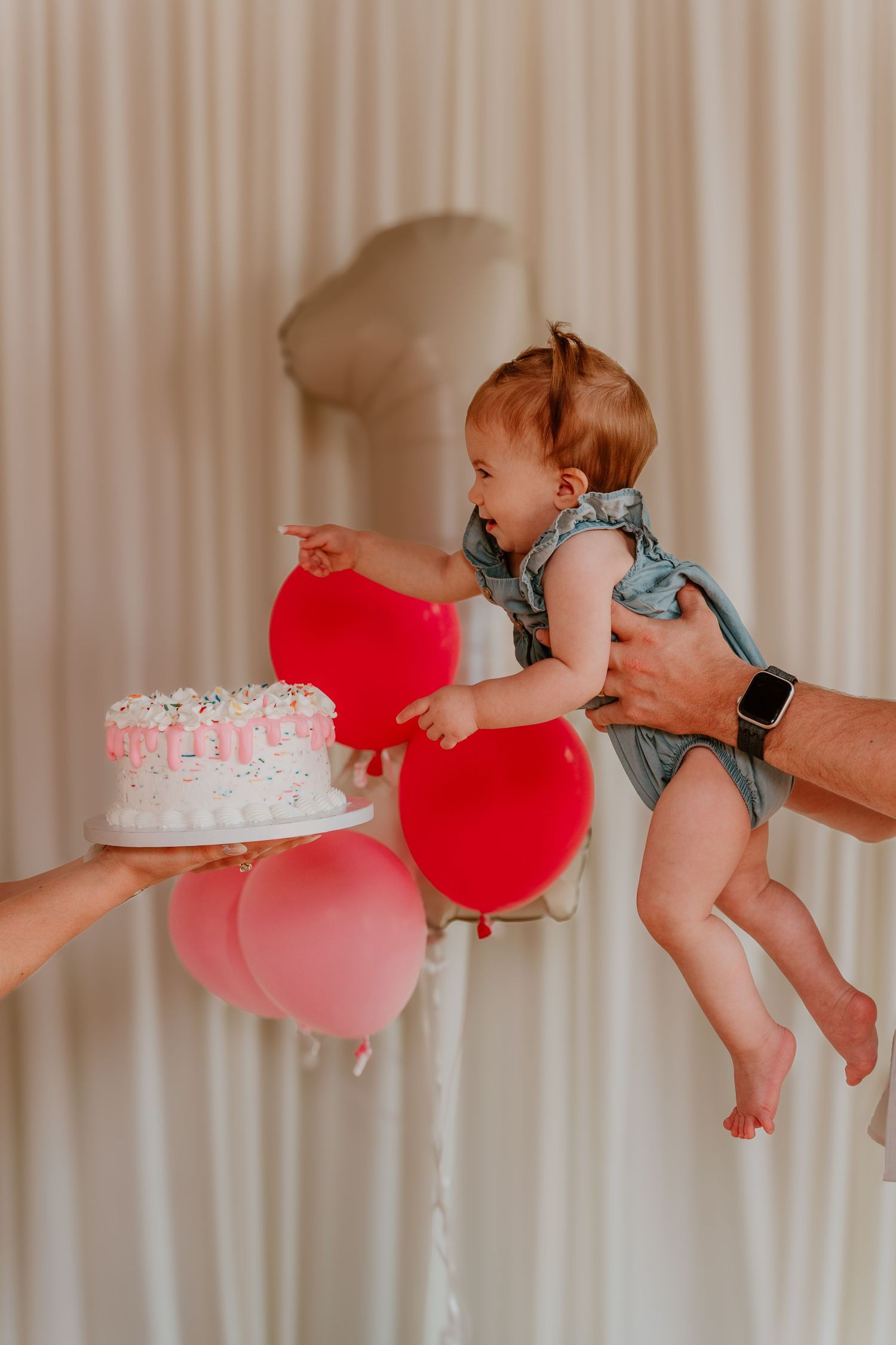 A baby reaching for a first birthday cake while being held, surrounded by red and pink balloons.