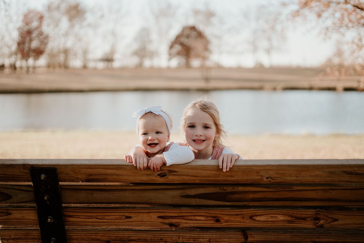 Two young girls smile over a wooden park bench near a serene lake on a sunny day.
