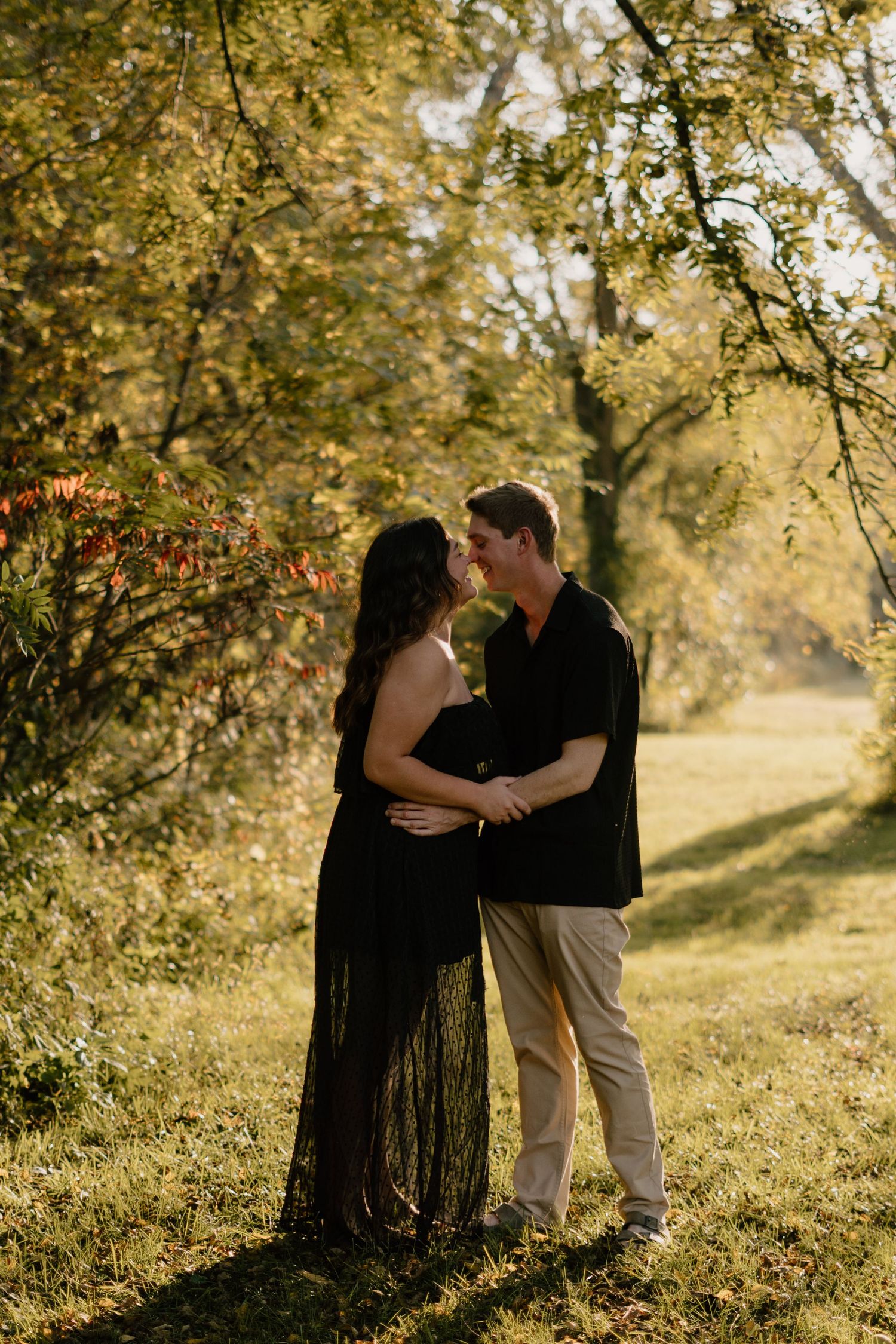 A couple shares a romantic moment outdoors, surrounded by golden autumn foliage, as the woman in a black gown cradles her baby bump.