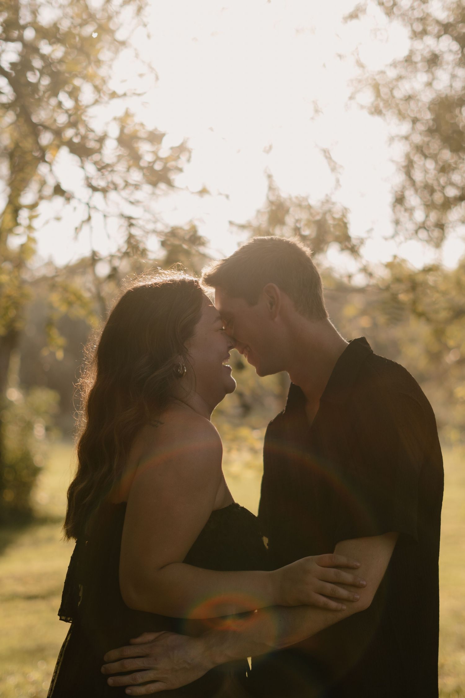 A couple in black outfits embracing and leaning in for a kiss in a sunlit park with golden backlight.