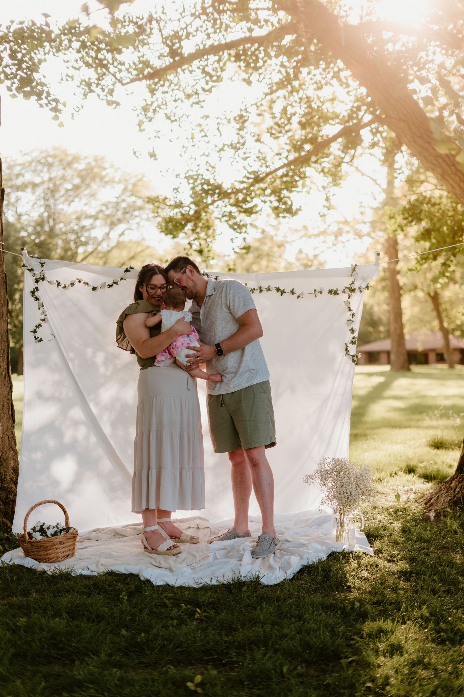 A couple shares a tender moment holding their newborn baby outdoors under a white fabric backdrop in golden sunlight.