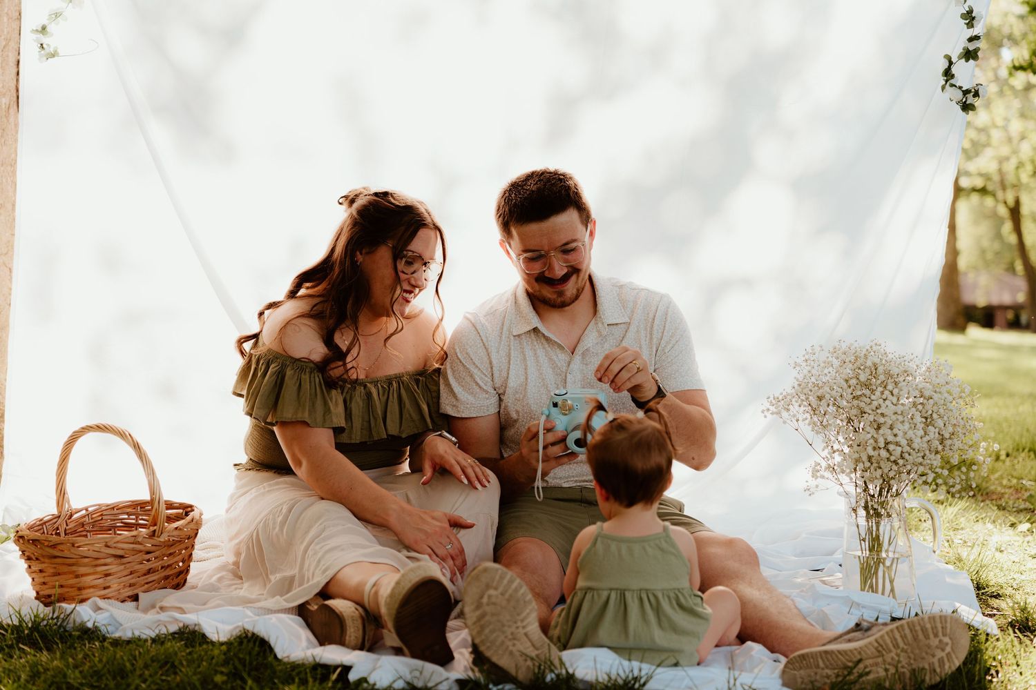 Happy young family enjoying a sunny outdoor picnic on a white blanket with a wicker basket and wildflowers.