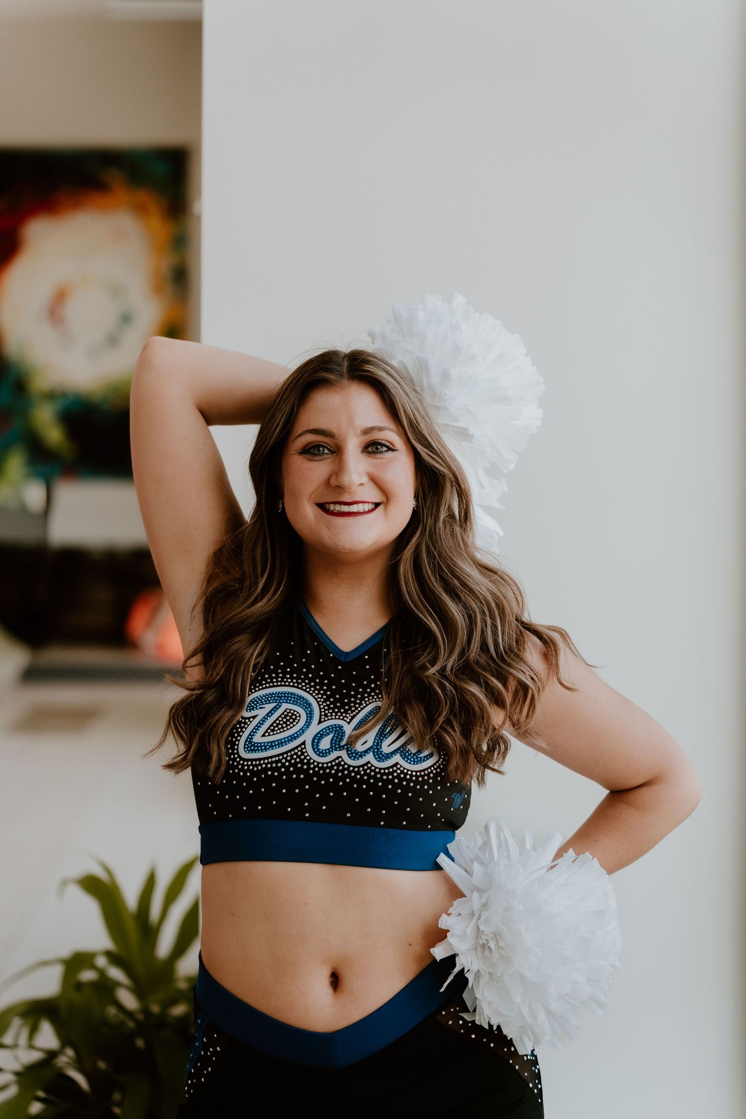 Cheerleader in black and blue Dolls uniform smiling with white pom poms in a bright indoor setting.