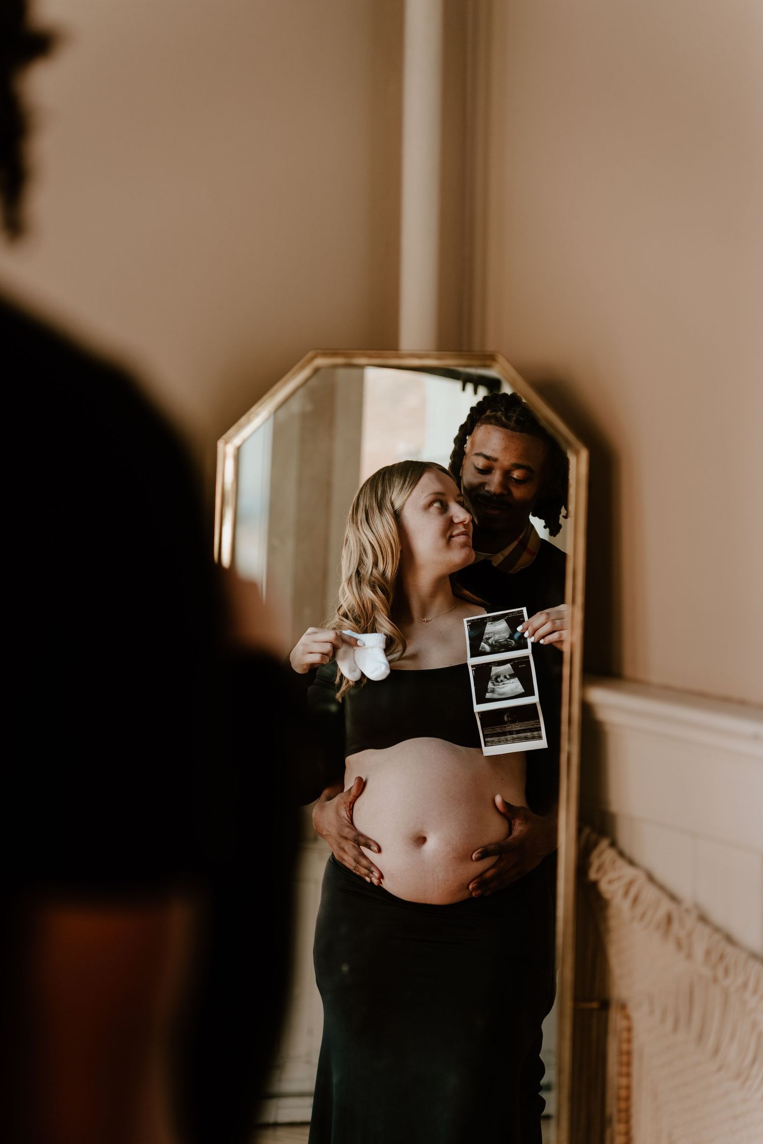 Pregnant couple holding ultrasound photos reflected in a gold mirror, celebrating their pregnancy together.