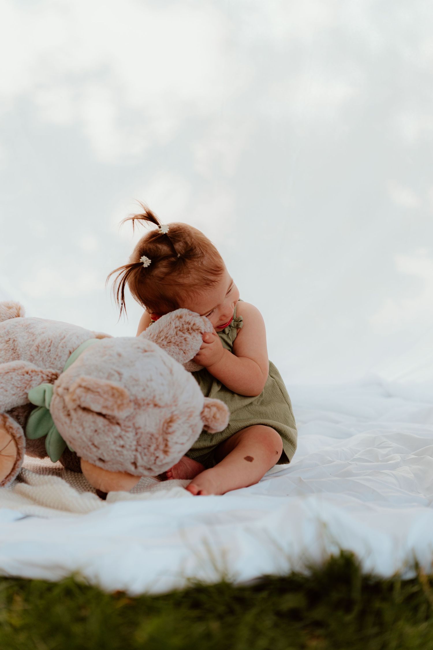 A toddler girl in a green romper sits on a white blanket outdoors, hugging a large teddy bear.