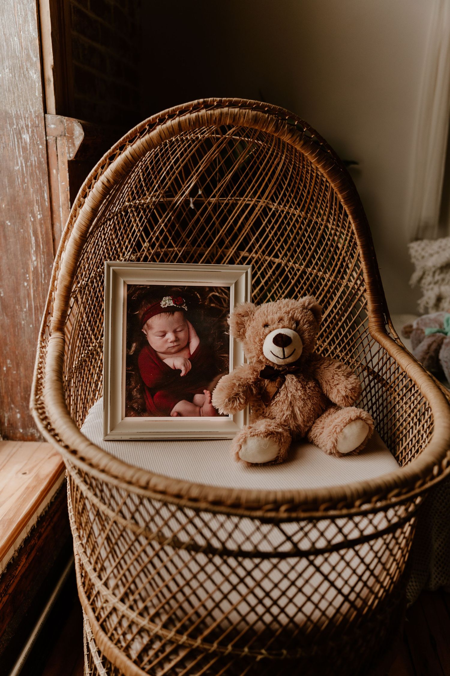 Wicker bassinet with a framed newborn baby photo and a brown teddy bear, styled in a warm vintage nursery setting.