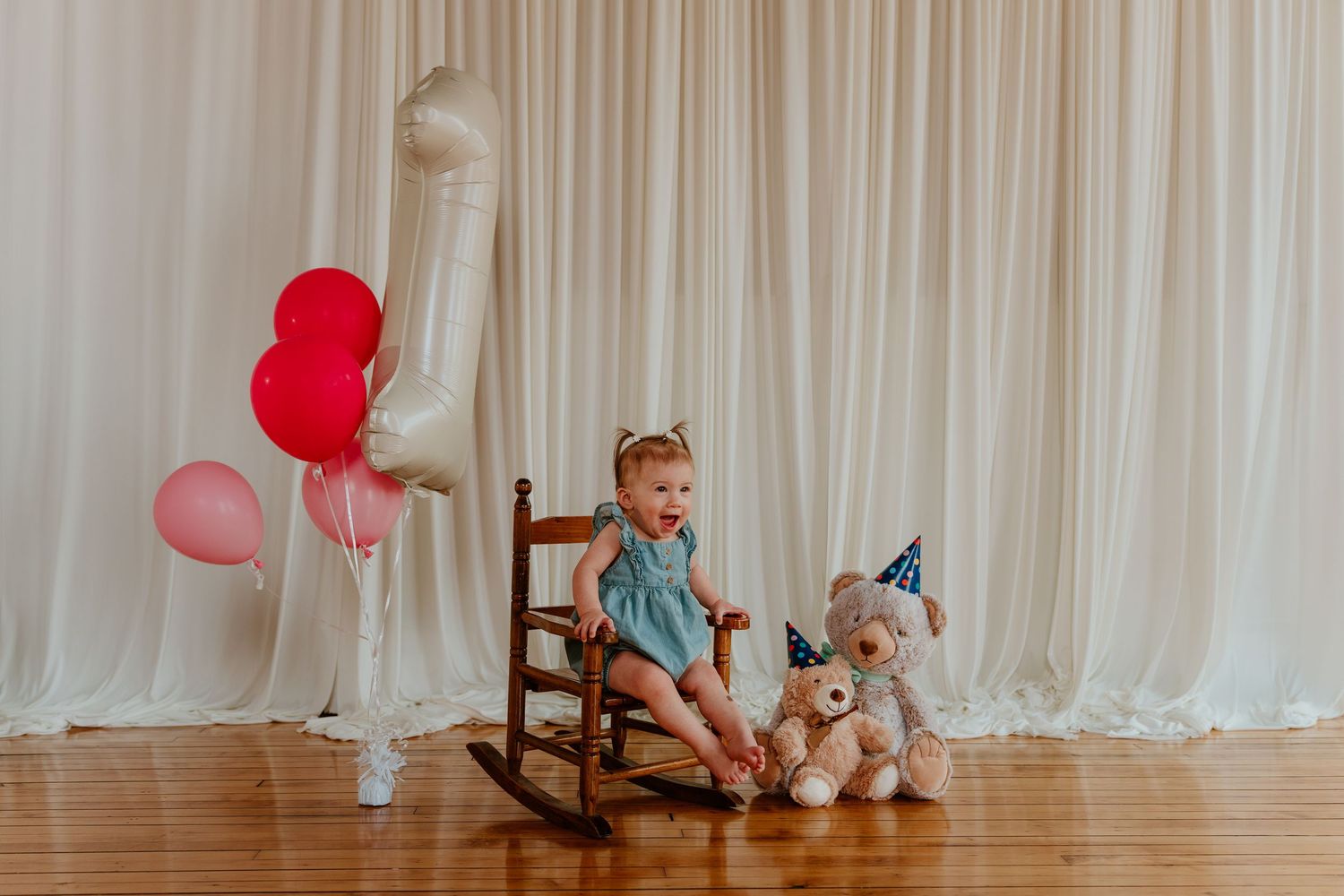 Toddler girl on rocking chair with teddy bears and birthday balloons celebrating first birthday.