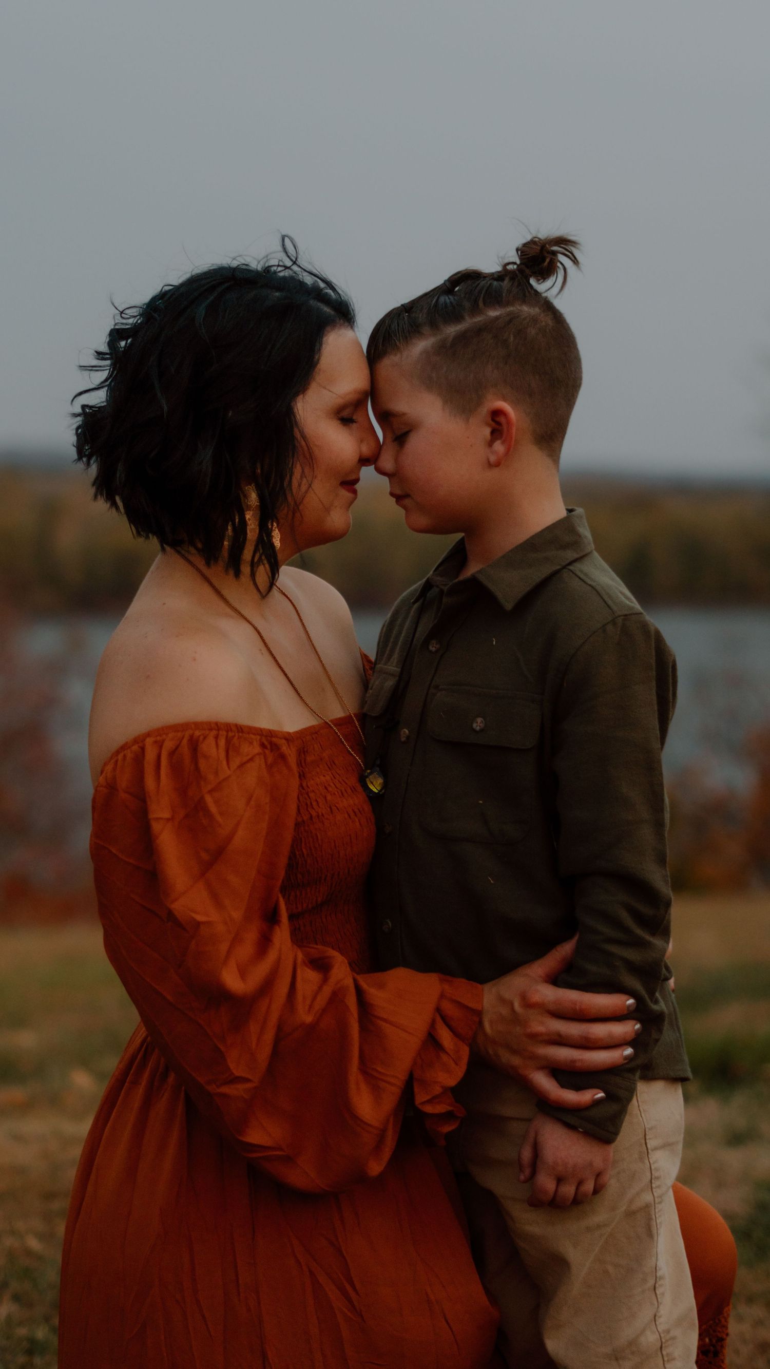Mother in orange off-shoulder dress touching foreheads with her young son in an outdoor autumn setting.