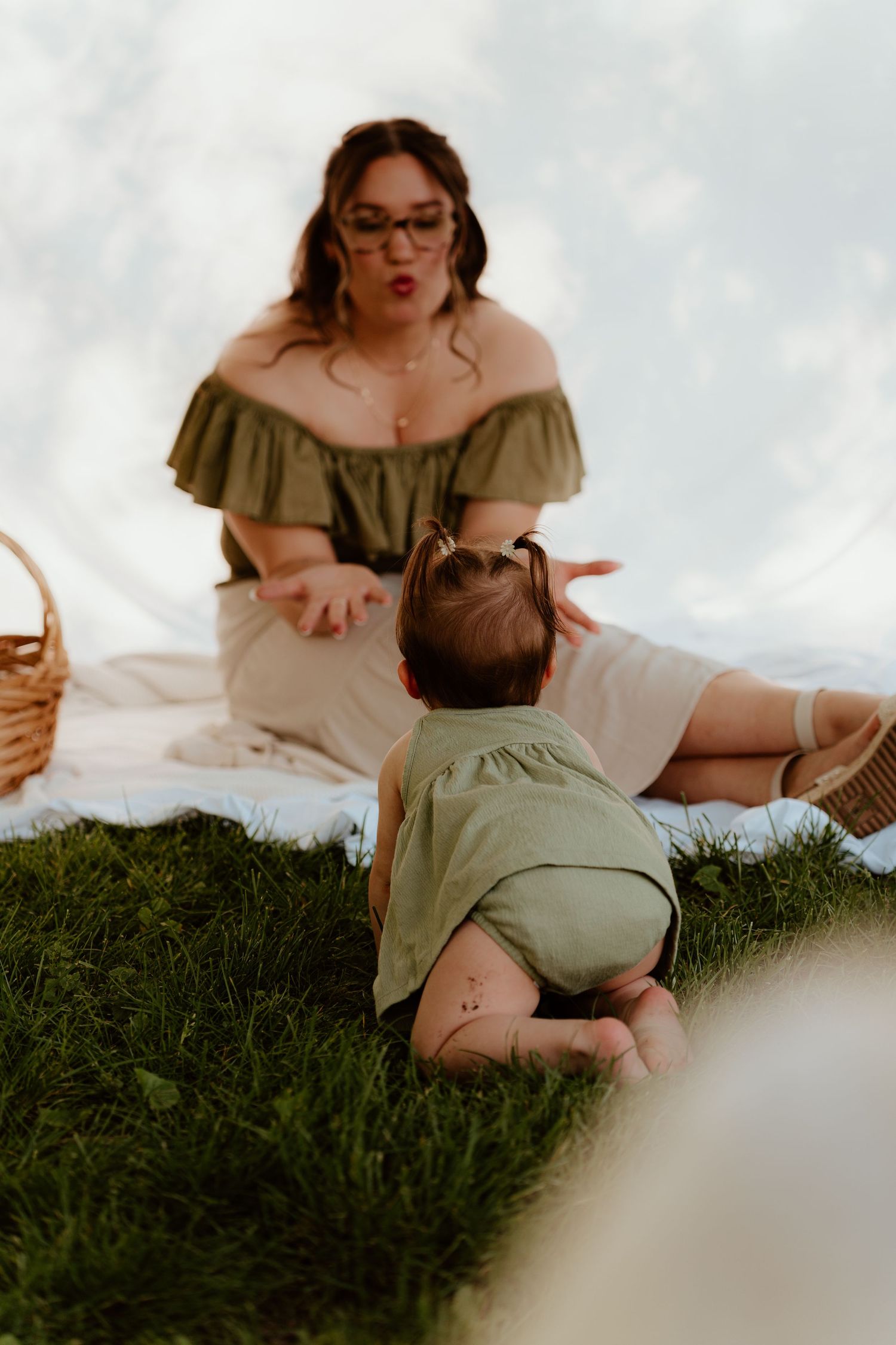 A mother in an off-shoulder green top sits on a blanket outdoors, engaging with her crawling baby on green grass.