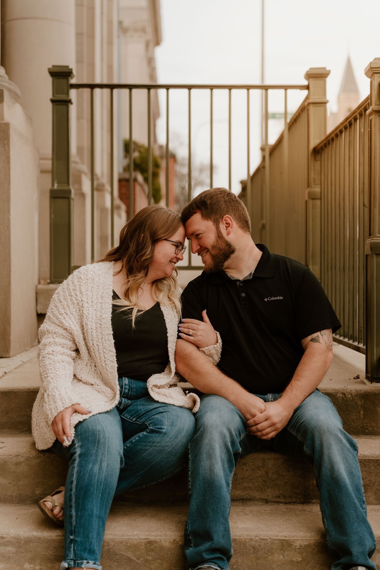 A couple sits on outdoor steps, leaning foreheads together and smiling, wearing jeans and casual tops in a warm urban setting.