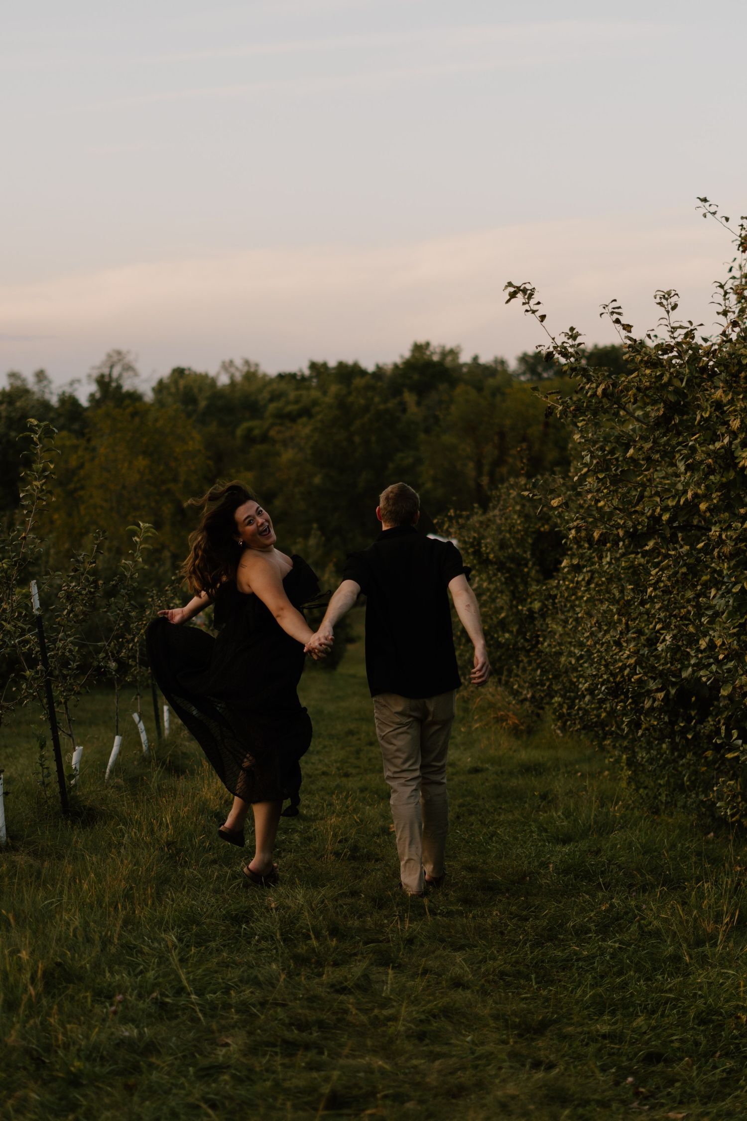 Couple holding hands running through orchard rows at dusk, woman in black dress laughing over shoulder.