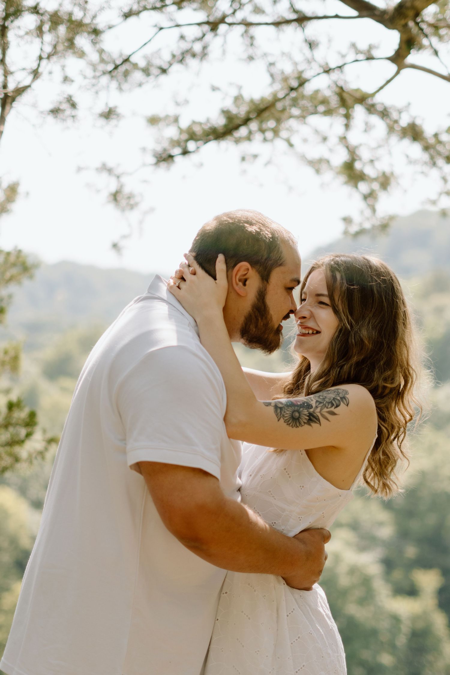 A smiling couple shares an intimate moment about to kiss under sunlit trees in nature.