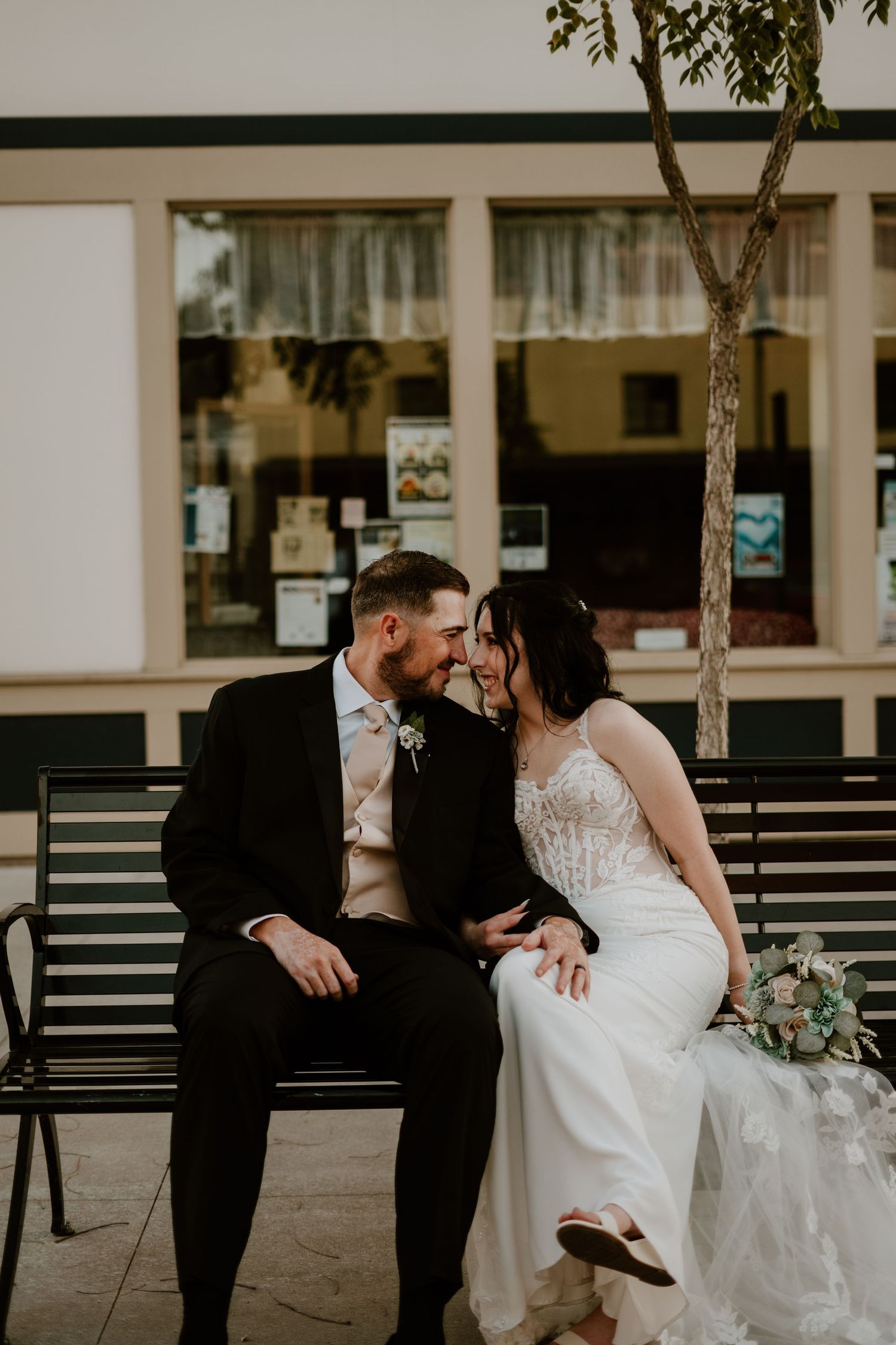 A bride and groom sit closely on a bench, smiling at each other, bride holding a bouquet, groom in black suit.
