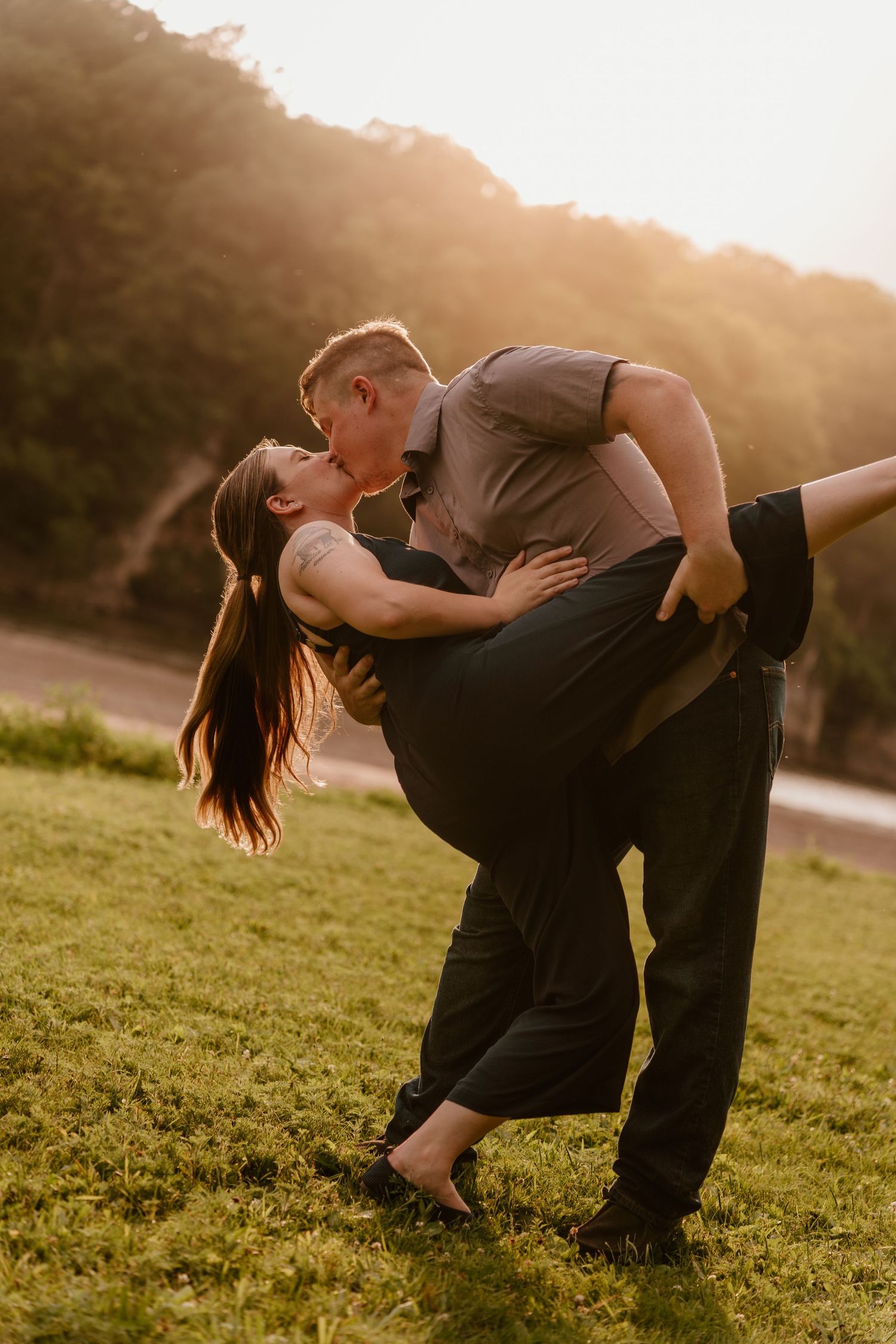 Couple sharing a romantic dip kiss at golden hour on a green lawn with sunlit hills in the background.