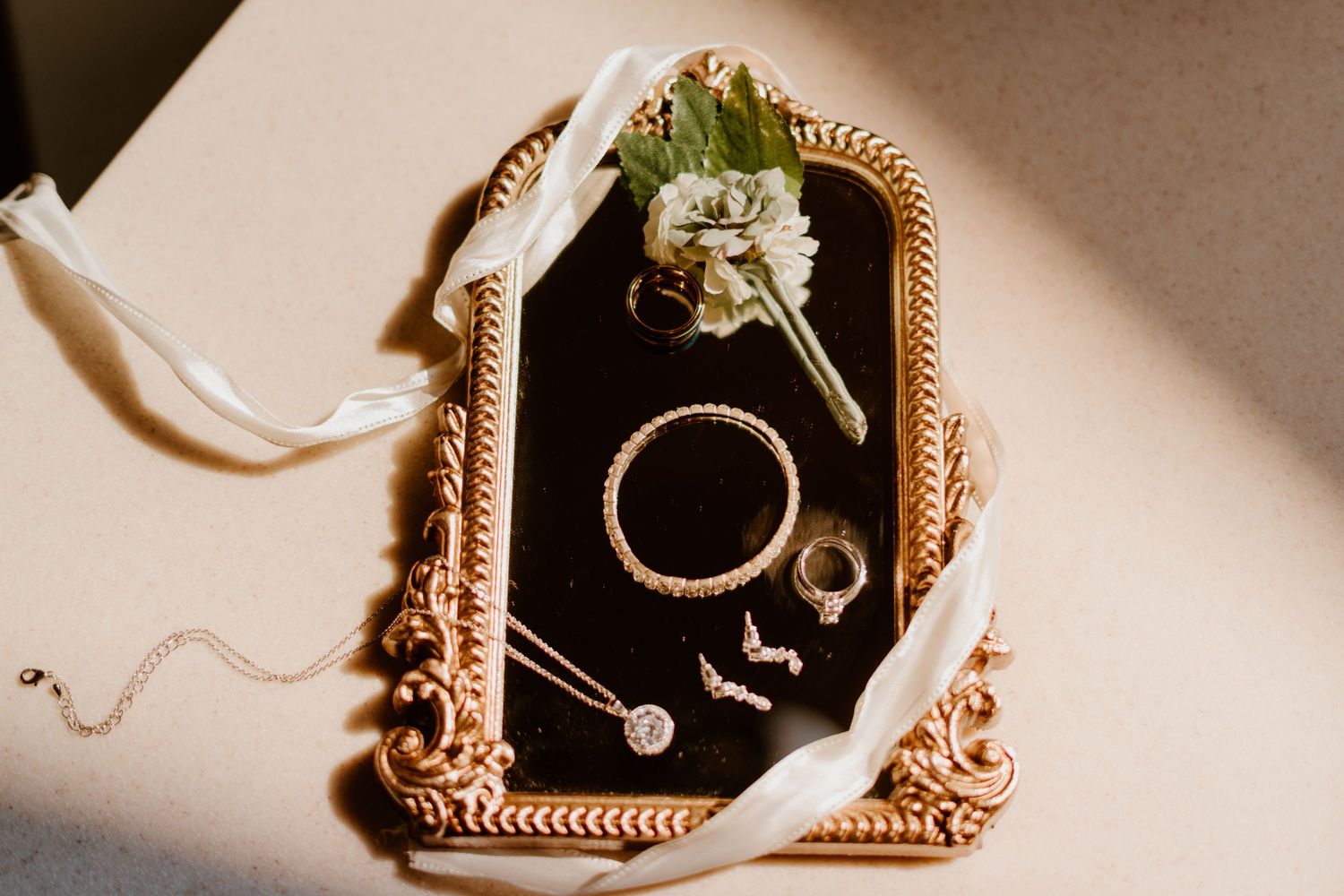 Gold ornate framed mirror displaying wedding jewelry including rings, necklace, and bracelet with white ribbon and floral boutonniere.