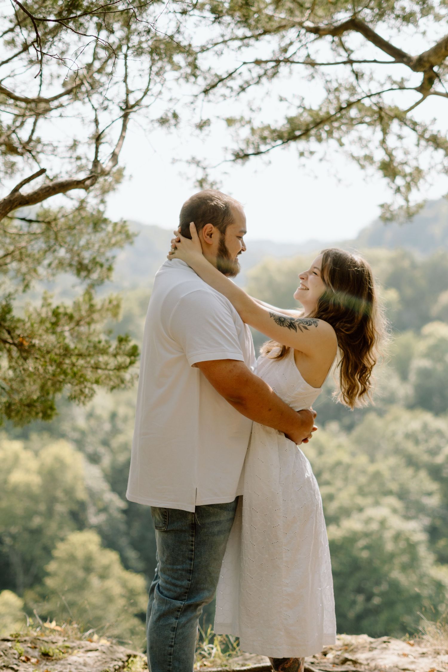 A couple embraces outdoors, smiling at each other with lush green forest hills in the background on a sunny day.