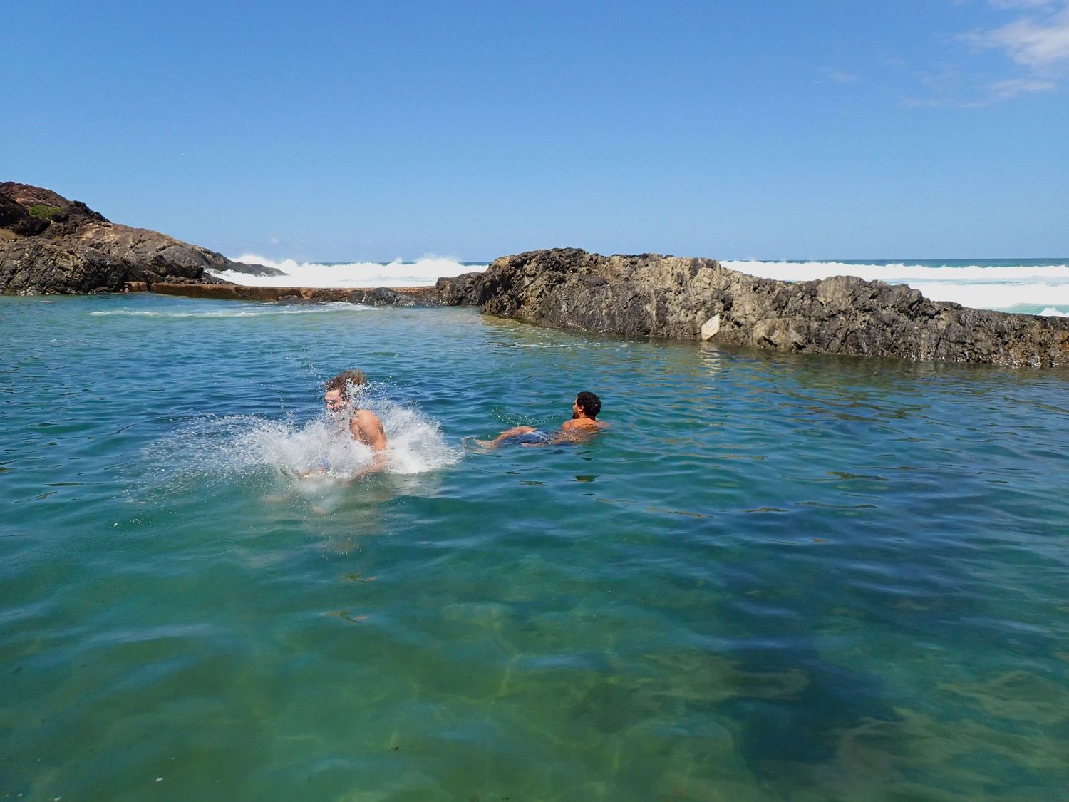 People swimming in vibrant turquoise waters near a rocky coastline on a sunny day. Sawtell Headland Beach.