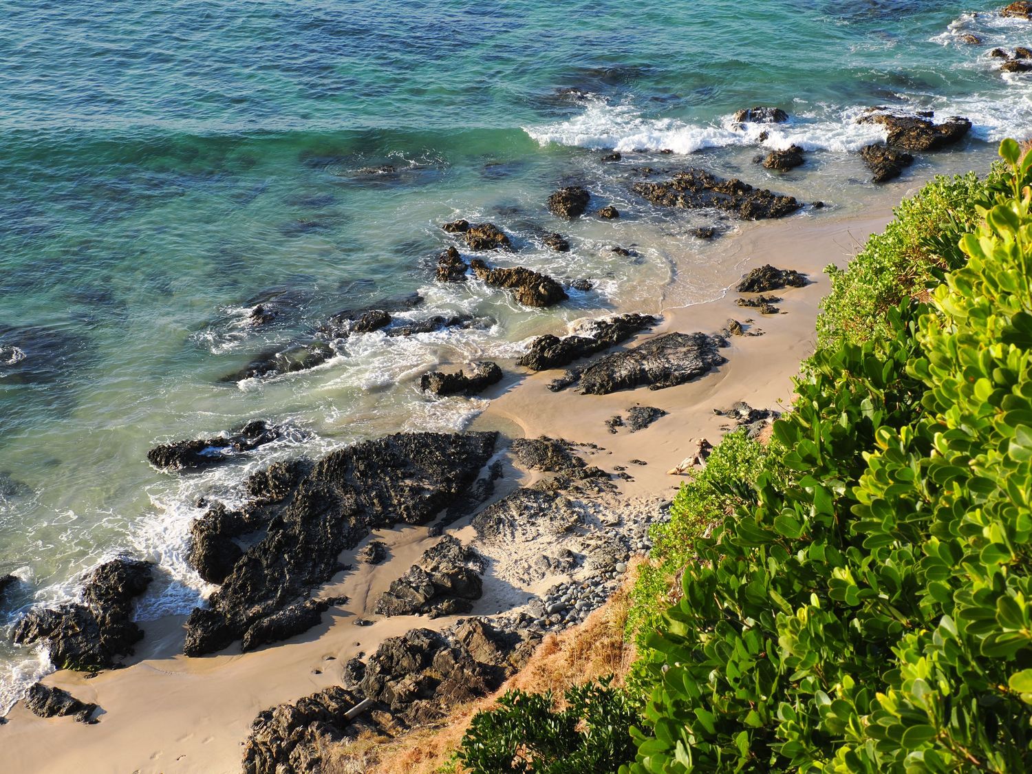 Turquoise ocean waves crash against black volcanic rocks along a sandy Byron Bay beach with lush green foliage.