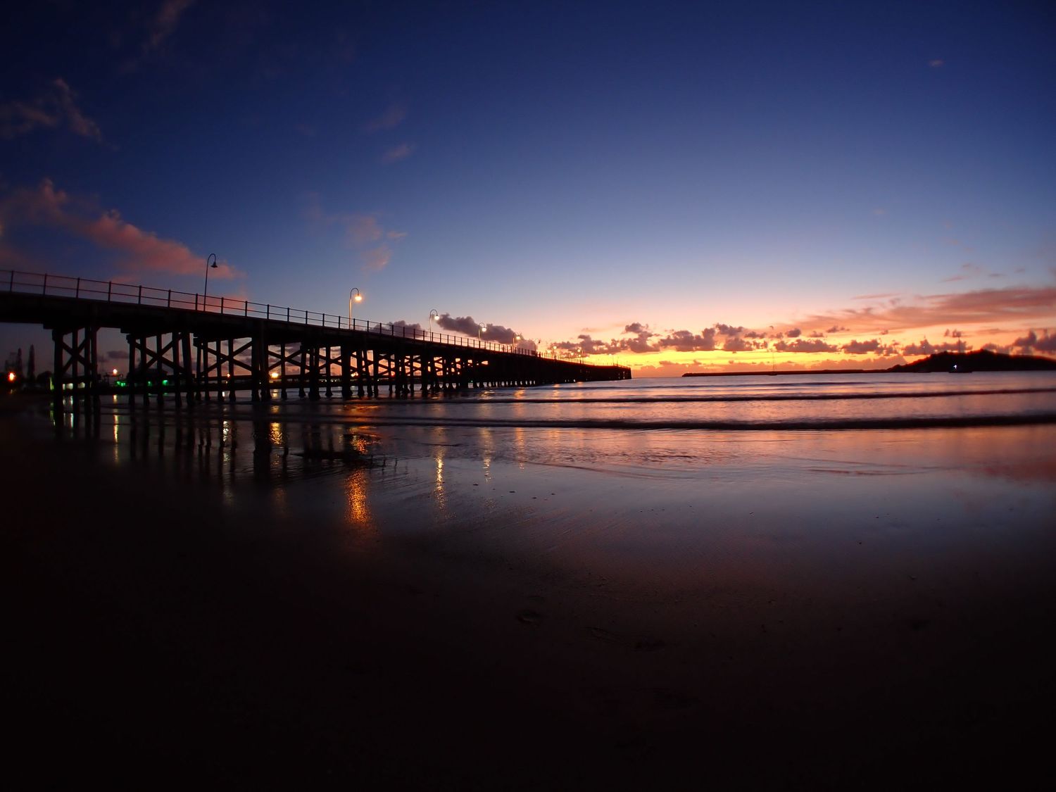 Panoramic view of a pier silhouetted against a vibrant sunset sky over calm waters. Coffs Harbour Jetty Sunsrise.