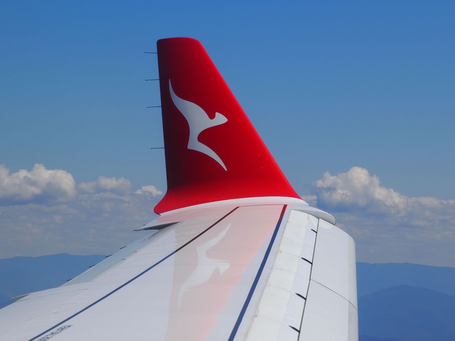 Red and white QANTAS airplane wingtip with blue sky and white clouds visible during daytime flight.