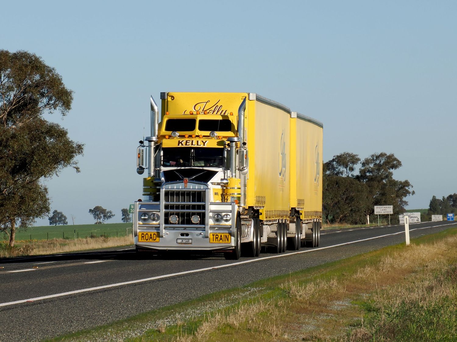Yellow Kenworth semi-truck hauling a trailer on a rural Australian highway surrounded by green fields and trees.