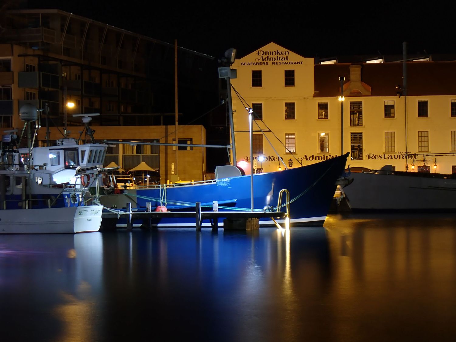 Illuminated boats and buildings reflect on calm harbor waters at night. Hobart Marina.