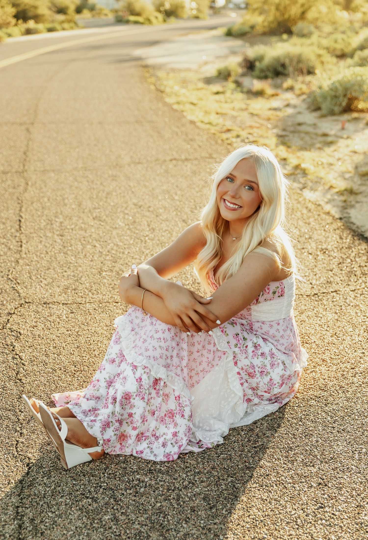 A person in a white floral dress sits on a sunlit path during golden hour creating a dreamy summer portrait.