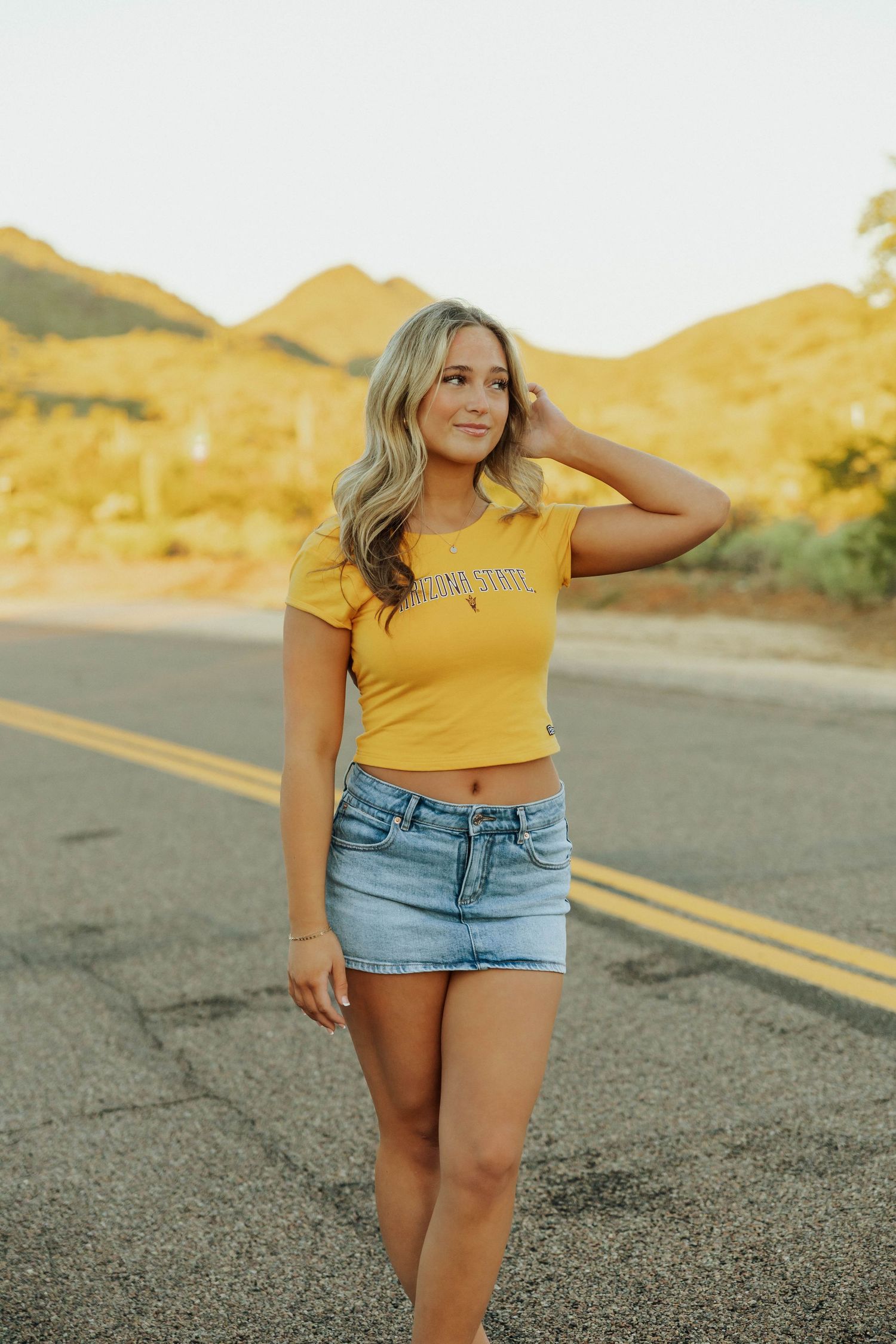 A person in a yellow crop top and denim shorts poses on an empty desert road at sunset.