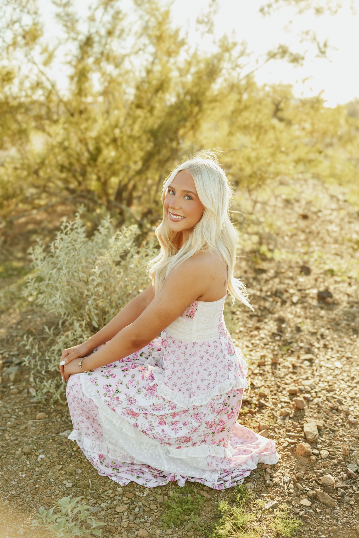 A person in a white and pink floral dress poses outdoors during golden hour in a desert landscape.