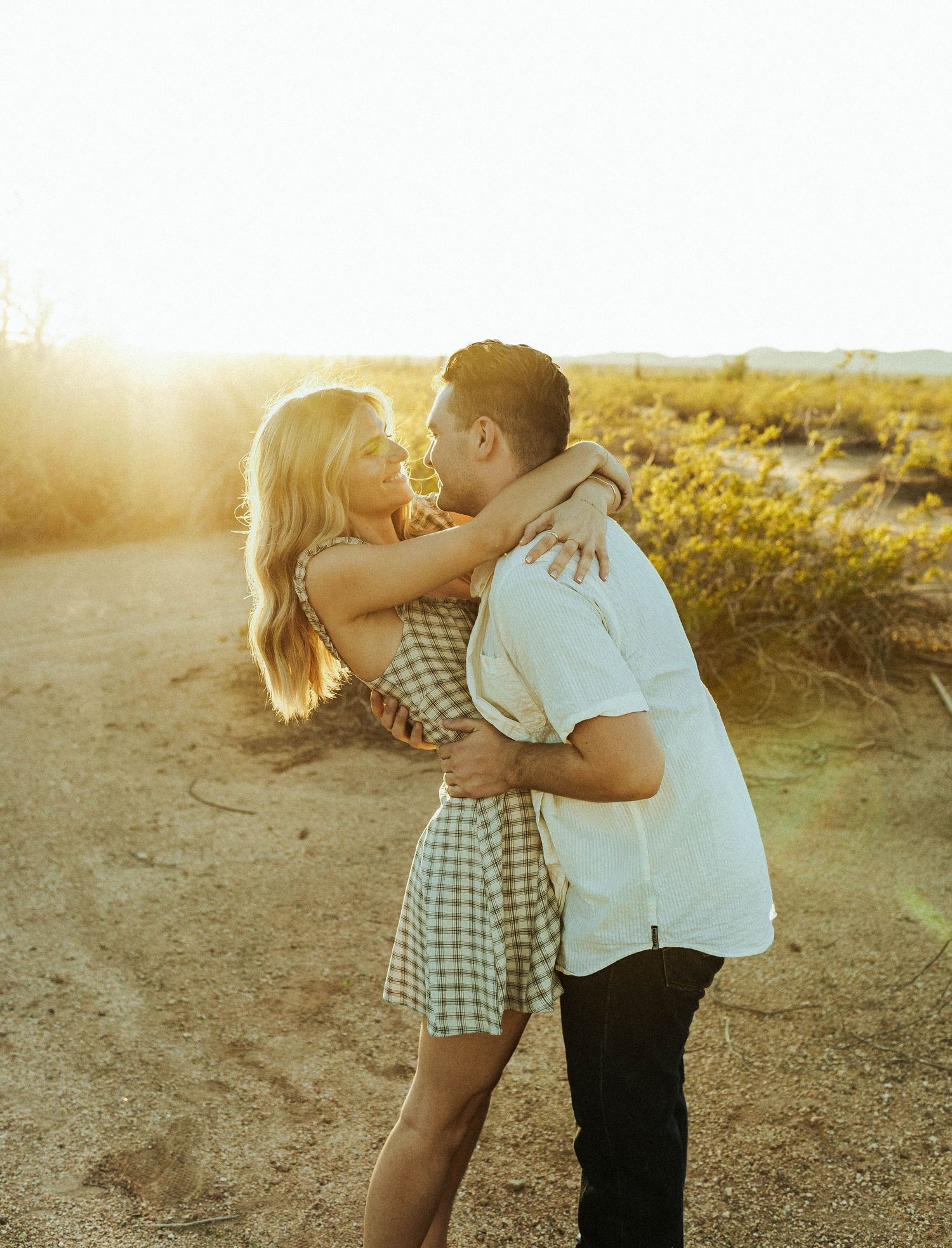 A couple embraces in a romantic moment at sunset on a dirt path surrounded by golden light and tall grass.