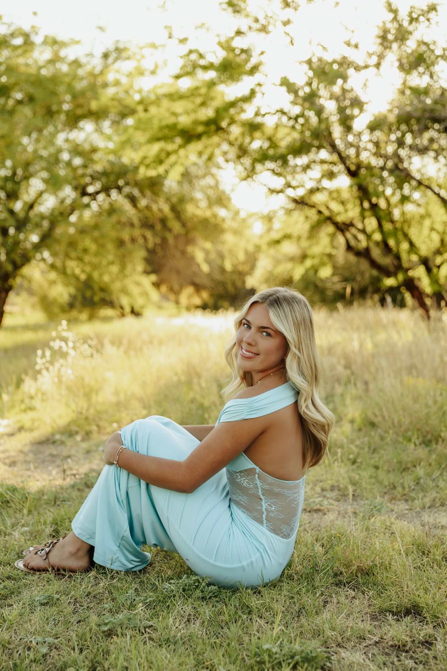 Smiling blonde woman in mint jumpsuit sitting on grass in a sunlit outdoor park setting.