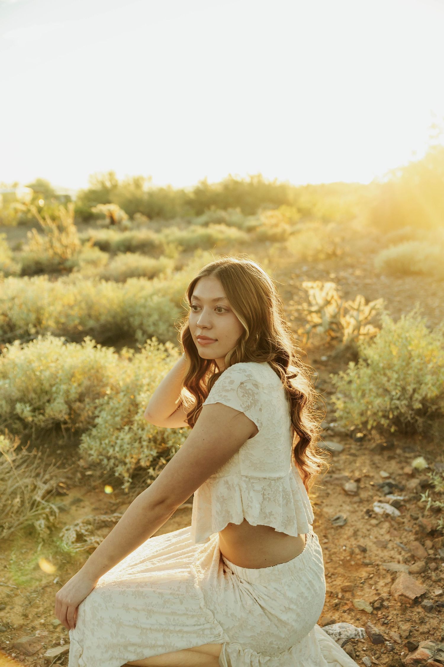 A person in a white lace dress poses gracefully on desert terrain during golden hour sunset.