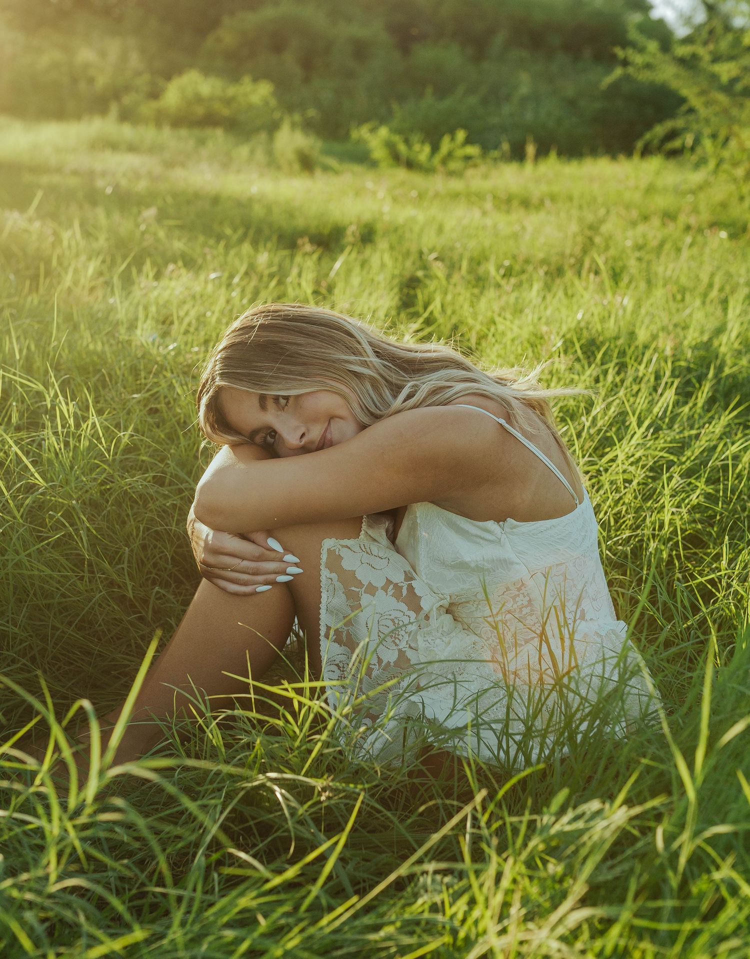 A person in a white dress sits peacefully in a sunlit meadow of tall grass during golden hour.