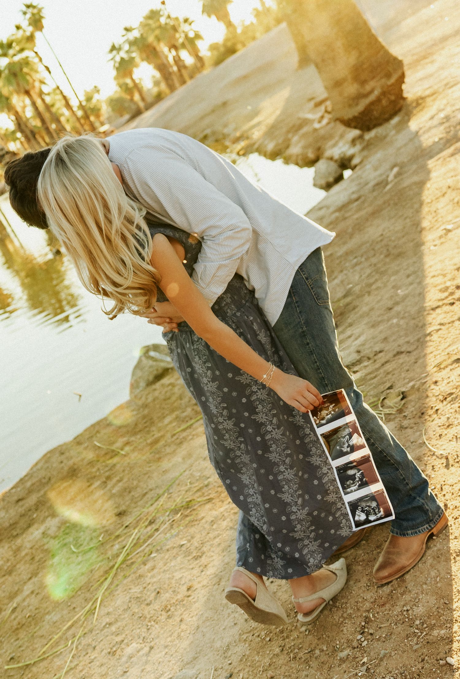 Someone in a white top and patterned skirt holds ultrasound photos while standing on a dirt path near a lake at sunset.