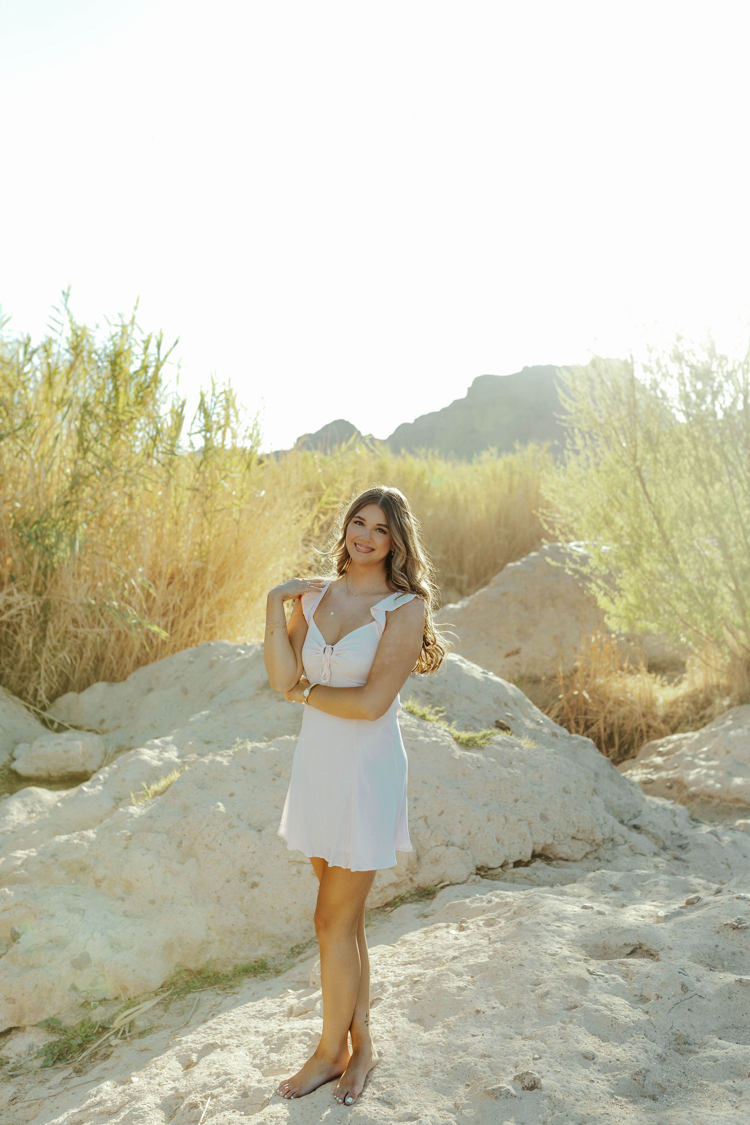 Desert senior photoshoot in white dress against golden brush and rocky terrain during sunset.