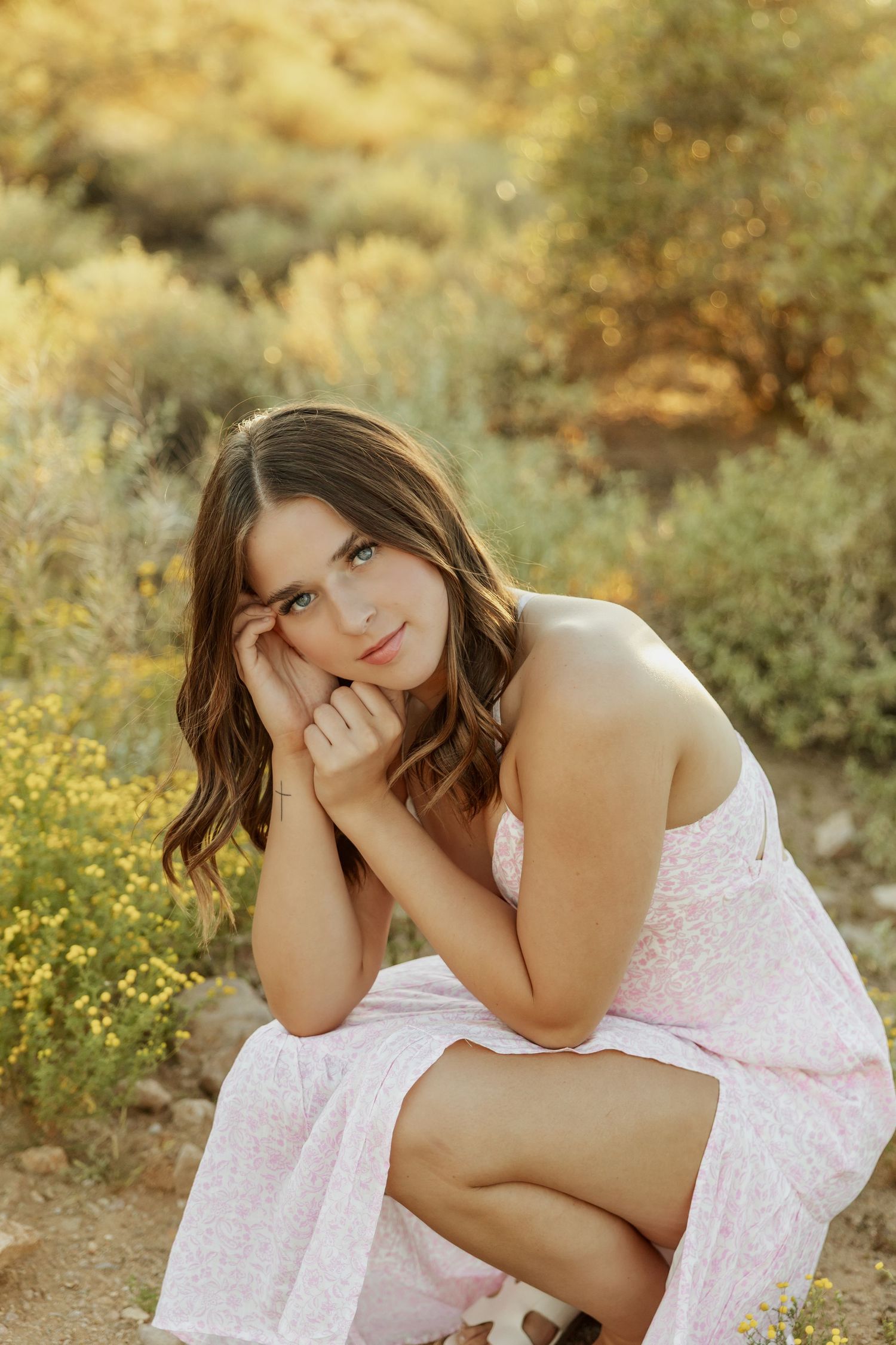 Young woman in pink dress crouching outdoors among wildflowers, resting face on hands in golden natural light.