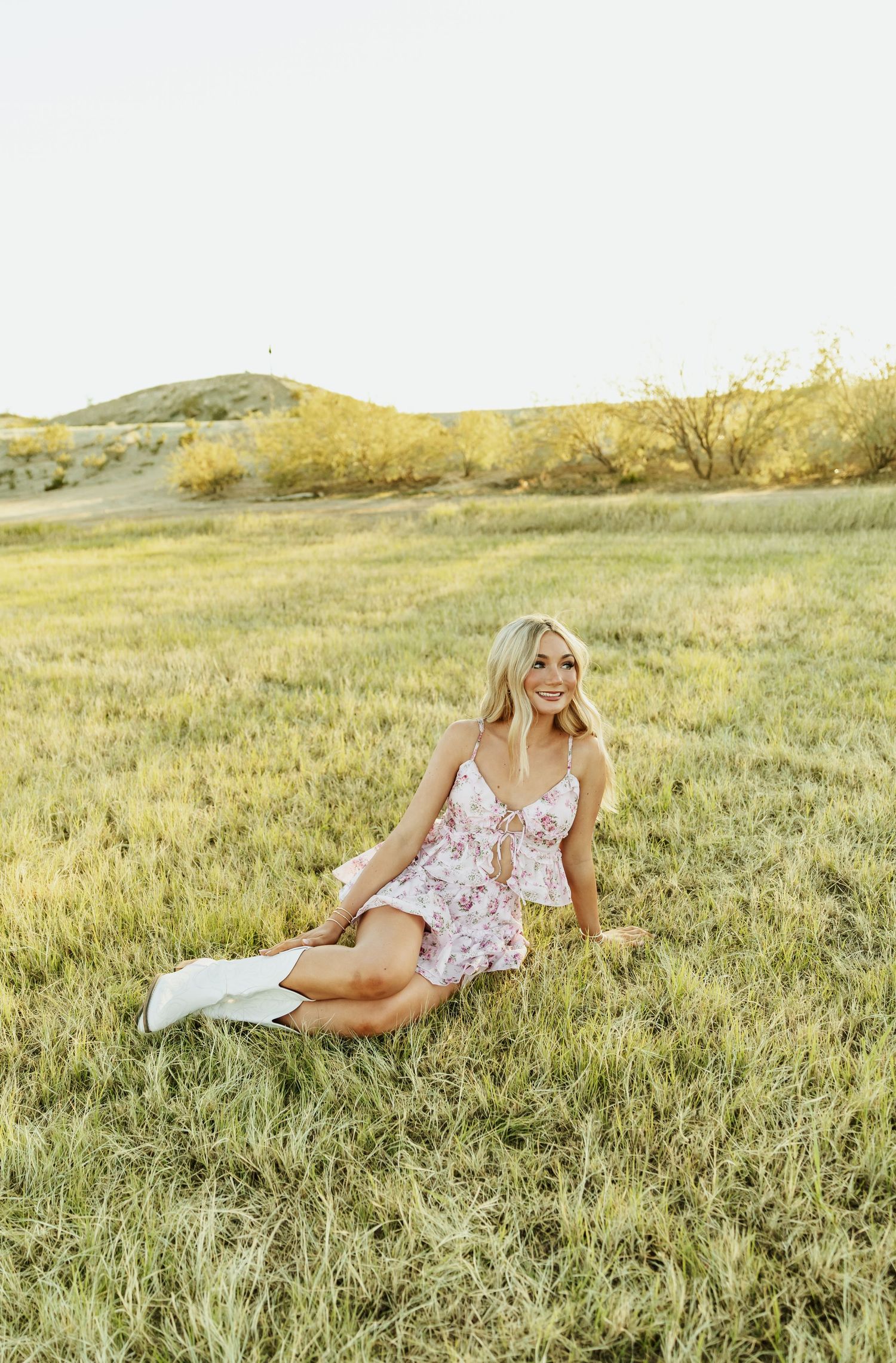 Blonde woman in floral mini dress and white boots sitting in green field with golden hour sunlight.