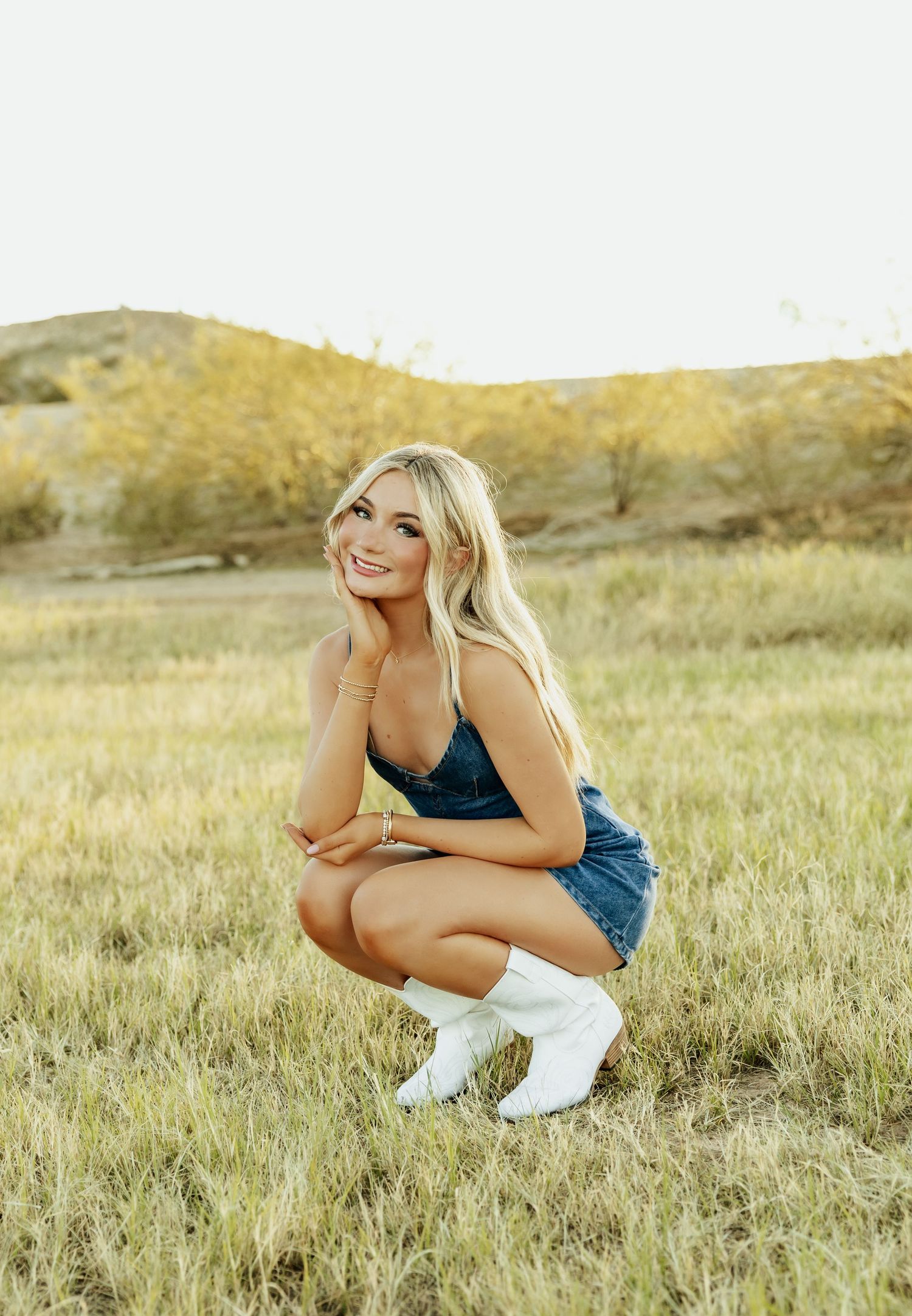 Blonde woman in denim romper and white cowboy boots crouching in a grassy field with hills in the background.