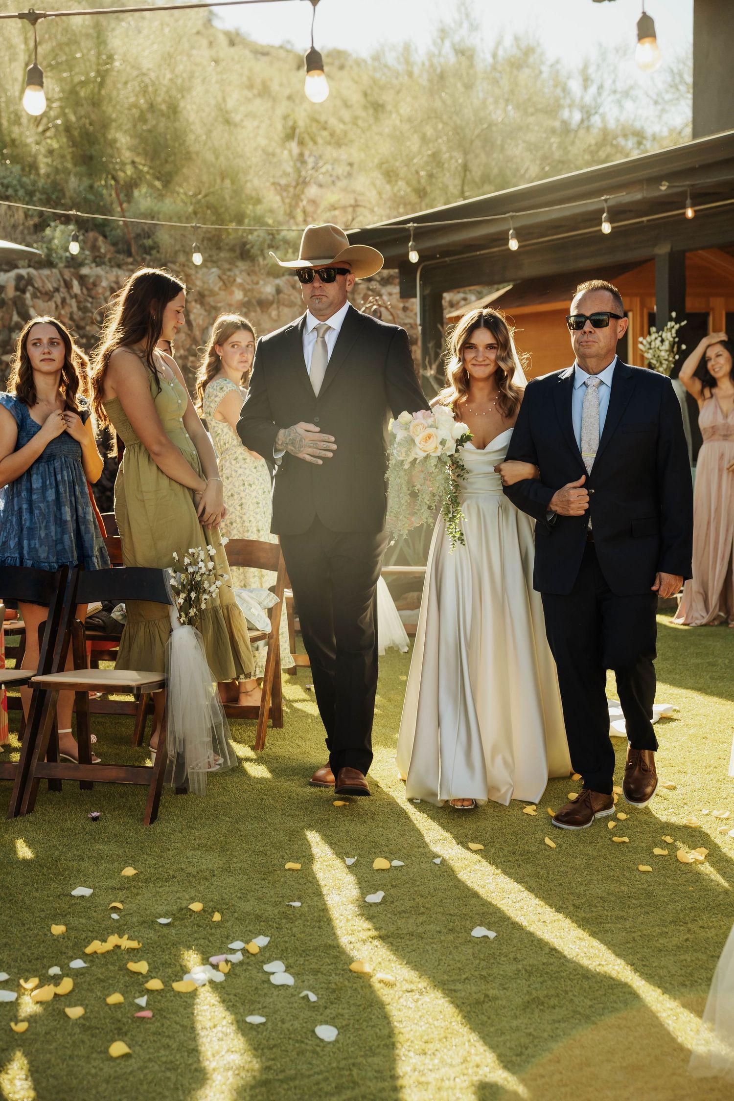 Wedding party mingles under string lights during golden hour at an outdoor reception.