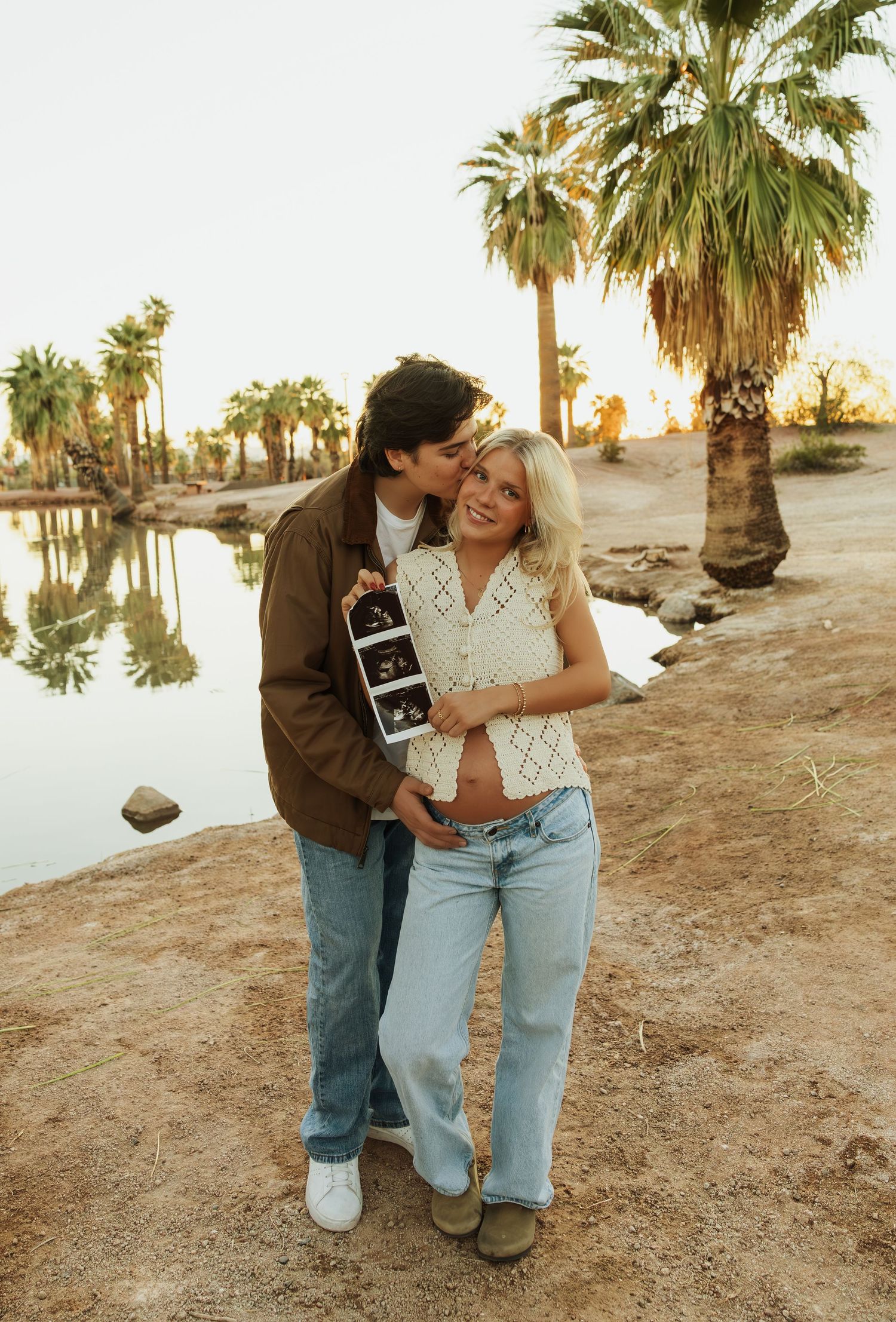 A couple shares a romantic moment at sunset near palm trees on a desert landscape.