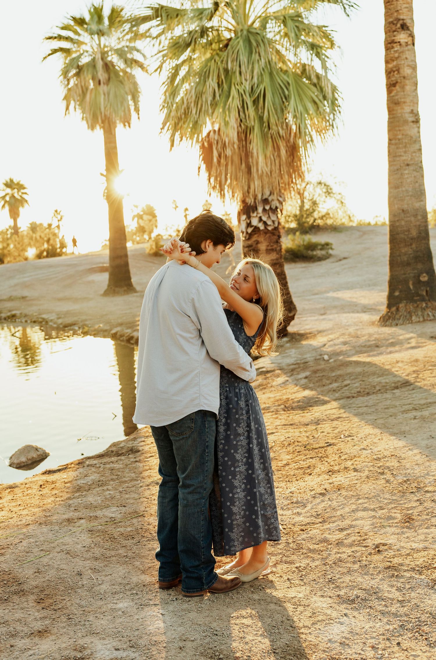 Couple embraces at sunset by palm trees and water's edge in warm golden light.
