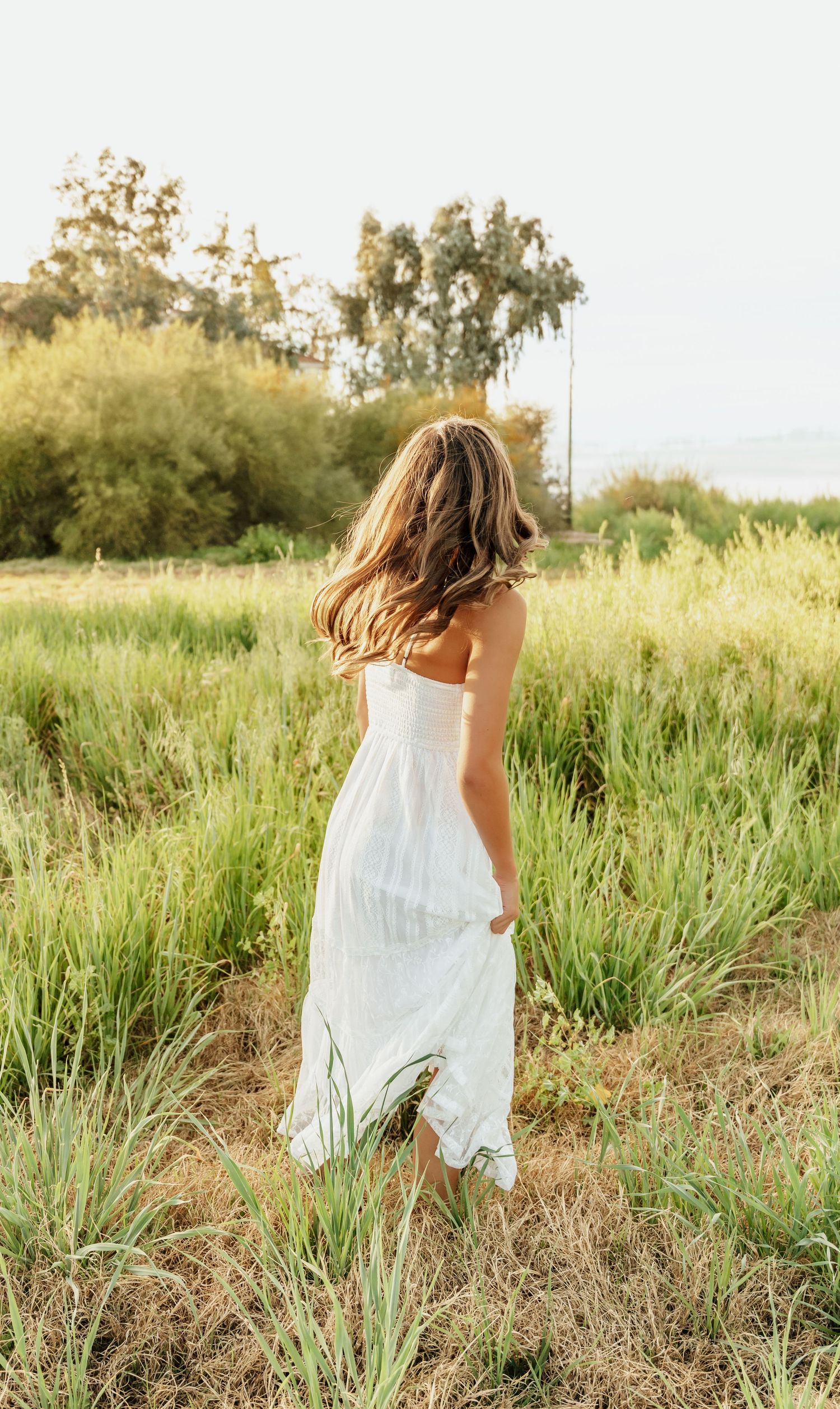 A white sundress flows in the breeze as someone walks through tall grass in a sunlit meadow at golden hour.