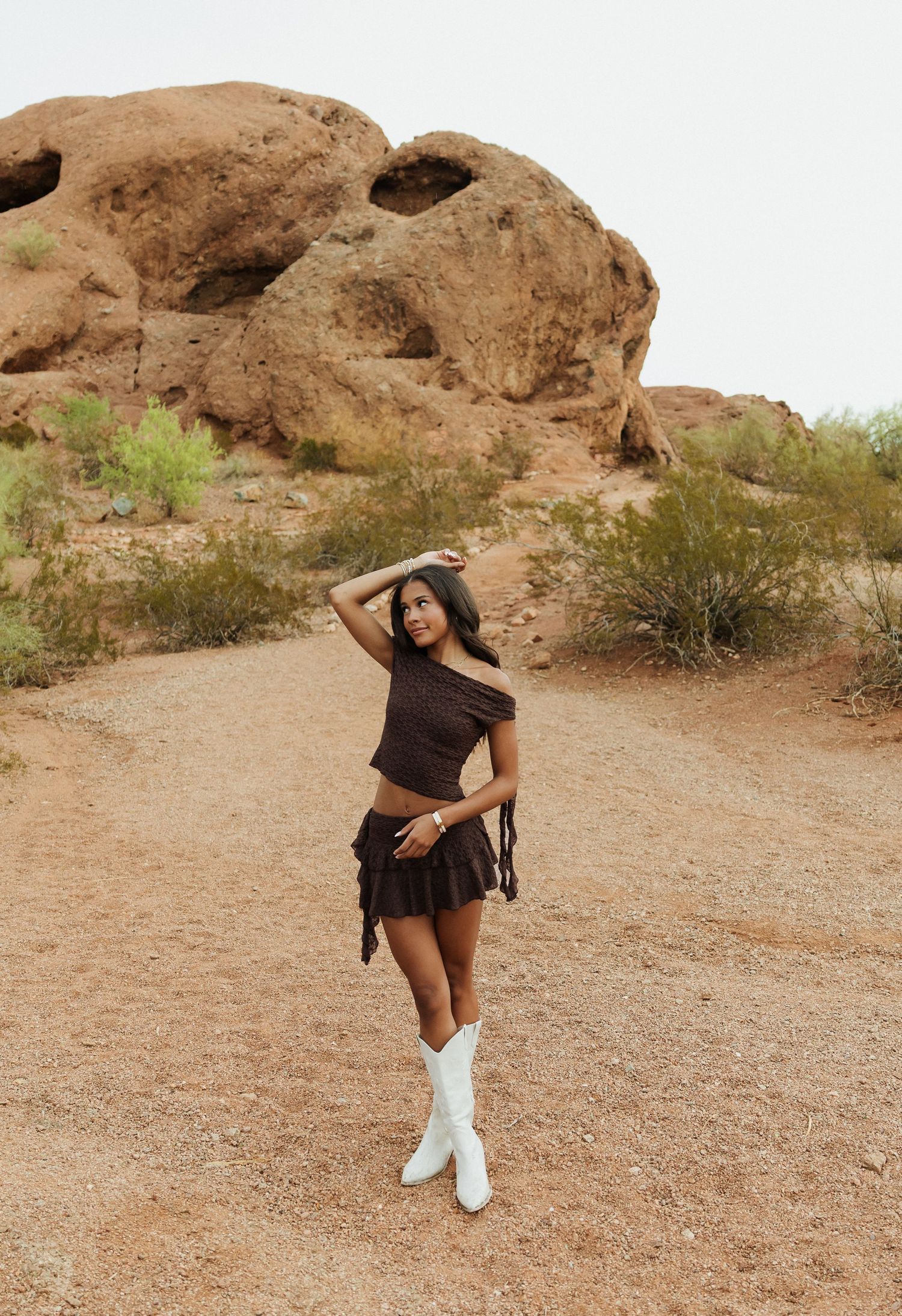 Woman in brown crop top, mini skirt, and white cowboy boots poses on desert trail with red rock formations behind her.