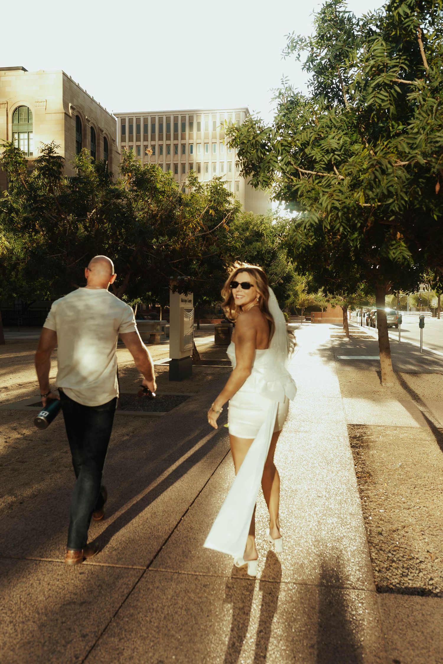 Couple strolling down a tree-lined sidewalk near modern buildings in warm sunlight. Elopement Photographer